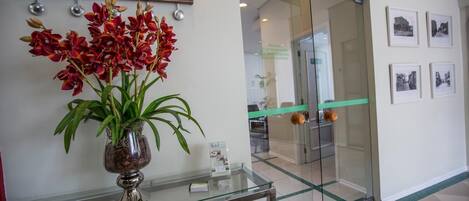 A reception area with a glass display case, a vase of red flowers, a clock with city names, and a door leading to a room with framed pictures.
