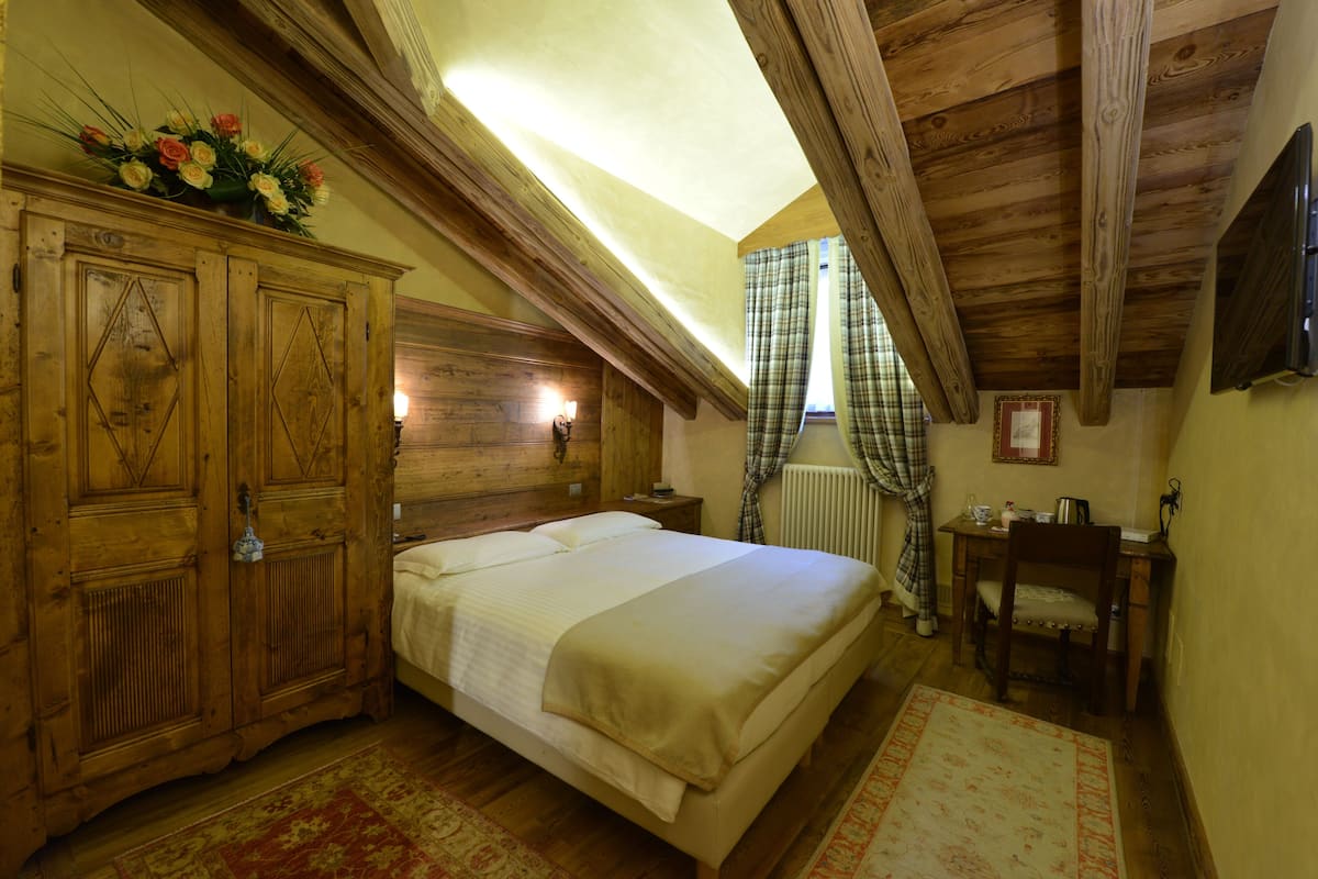 A bedroom with a wooden headboard, a bed with white linens, a wooden wardrobe, a small bedside table, a chair, and a window with curtains.