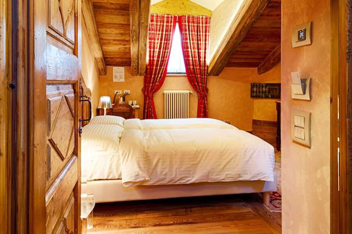 A wooden room with a bed, a window with red curtains, and a sloped ceiling.