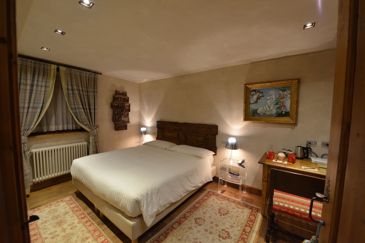 A bedroom with a large bed, bedside lamps, a wooden headboard, a radiator, a window with curtains, a painting on the wall, and a desk with a lamp and a red mug.