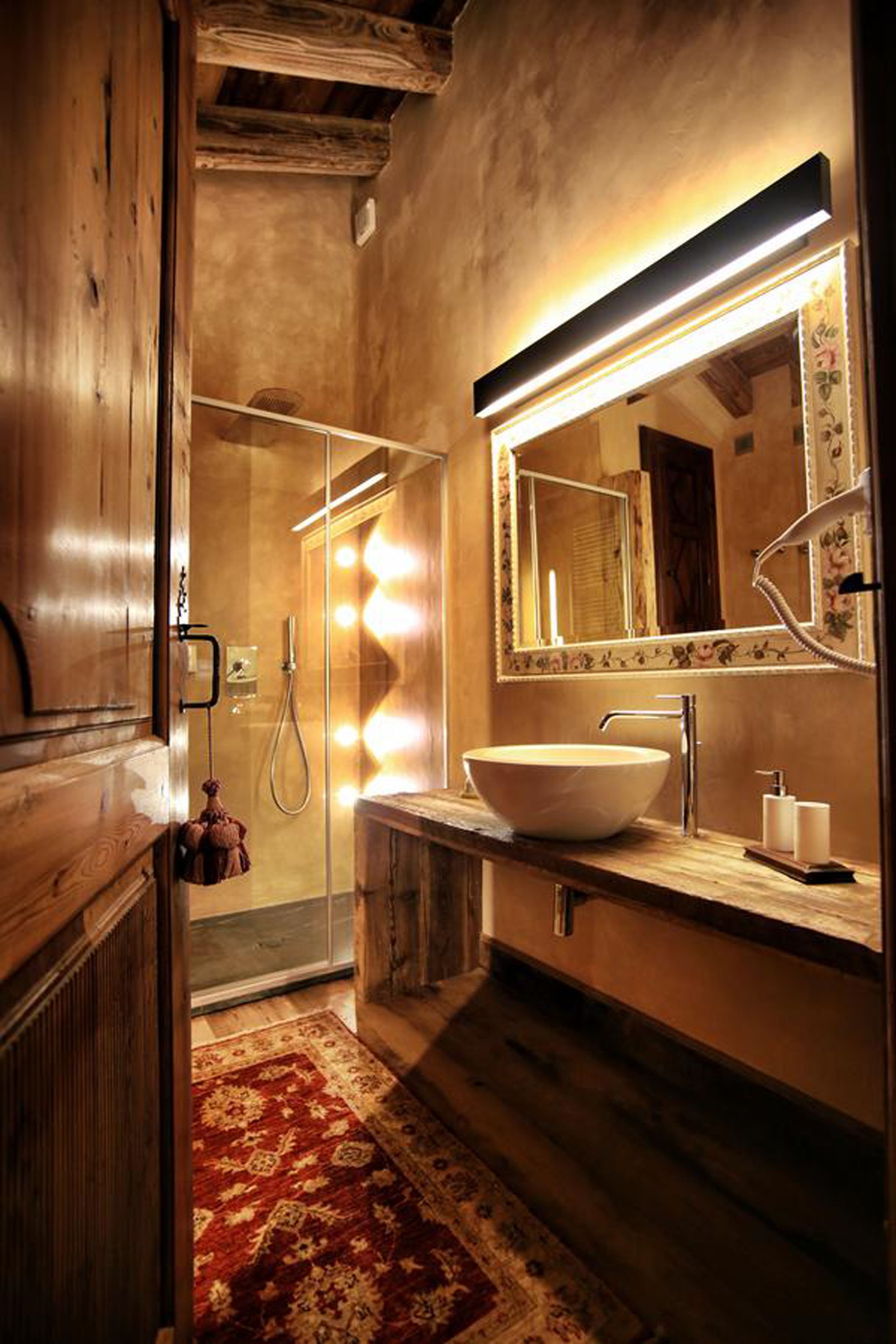 A bathroom with a wooden interior, a large mirror with lights, a white sink, and a shower.