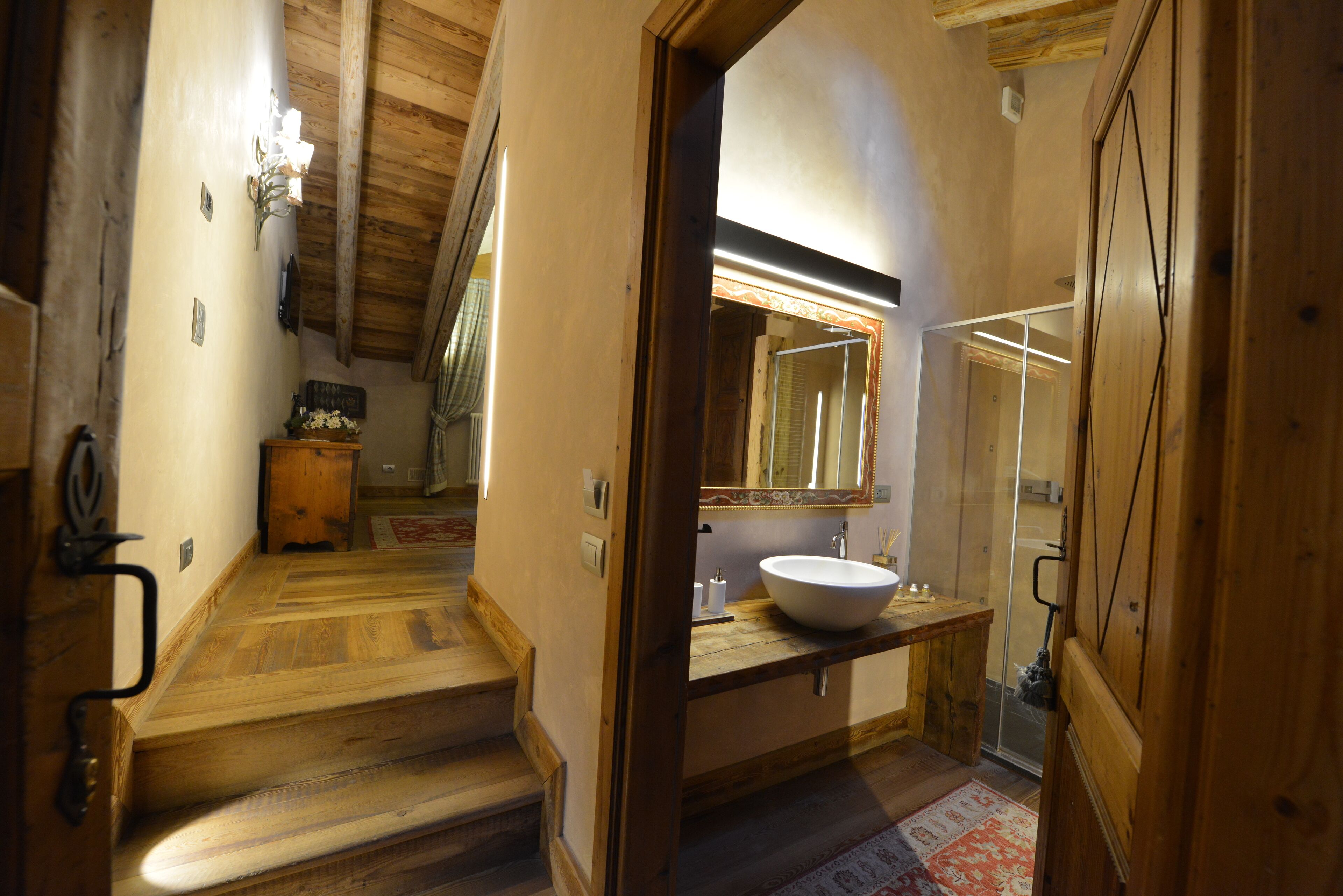 Un escalier en bois mène à une salle de bain équipée d’un grand miroir, d’un lavabo et d’un espace douche.