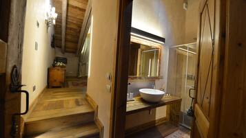 Un escalier en bois mène à une salle de bain équipée d’un grand miroir, d’un lavabo et d’un espace douche.