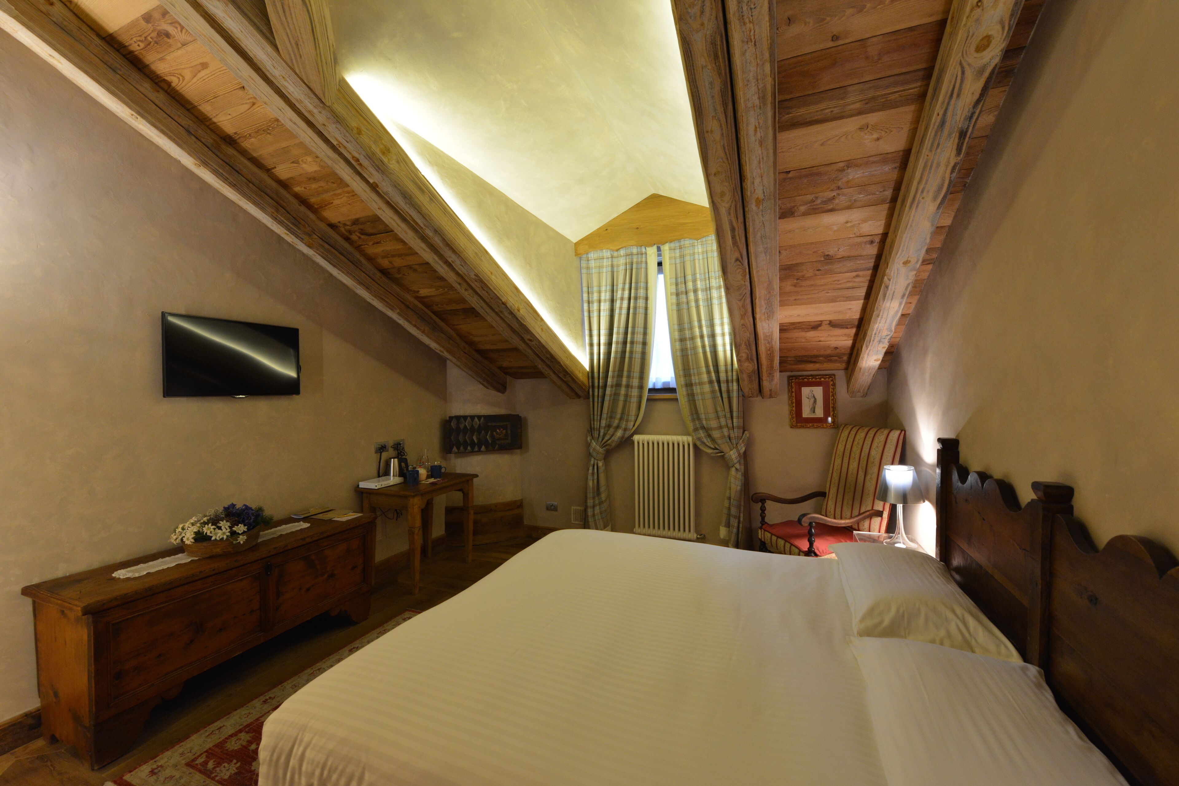 A bedroom with a large bed, wooden ceiling beams, a dresser, a chair, a table, and a television on the wall.