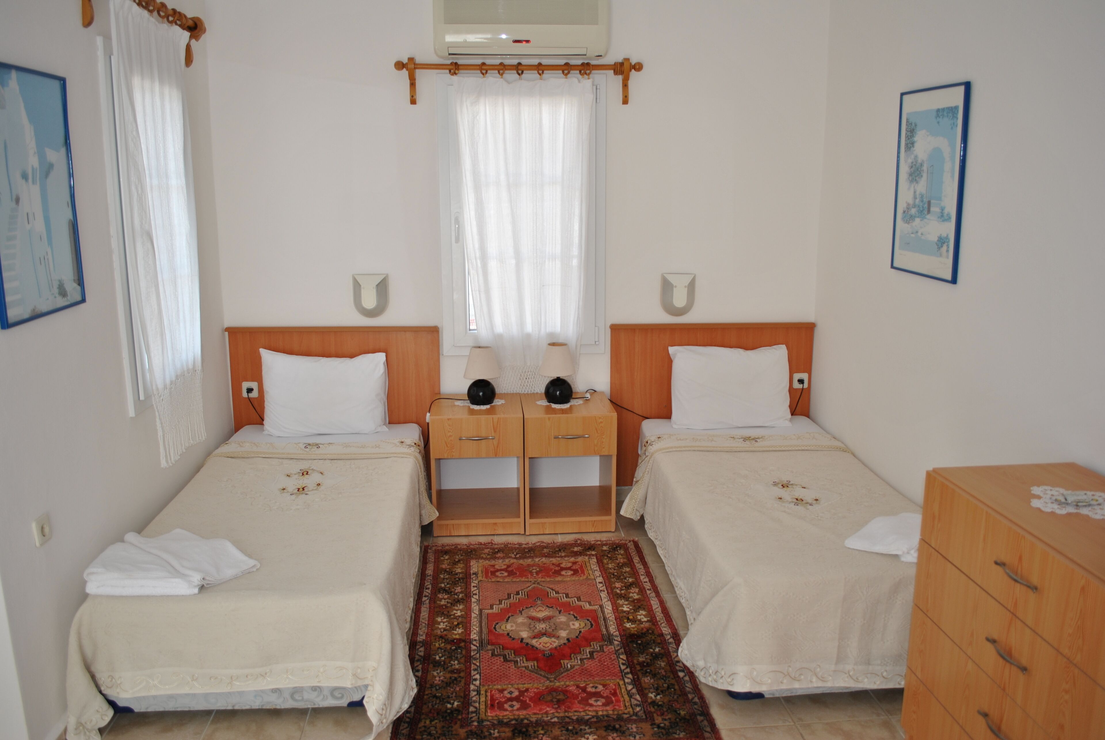 Une chambre double avec deux lits, une table de chevet en bois, un tapis rouge à motifs et une commode en bois.