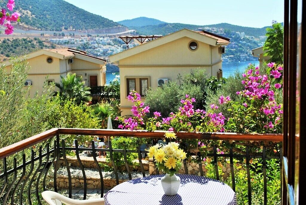A balcony with a table and chairs, overlooking a coastal area with houses and a view of the sea.