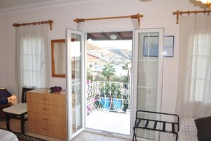 A bedroom with a wooden dresser, a chair, a bed, and a balcony with a view of a town and mountains. - Hotel Dionysia (Kas)