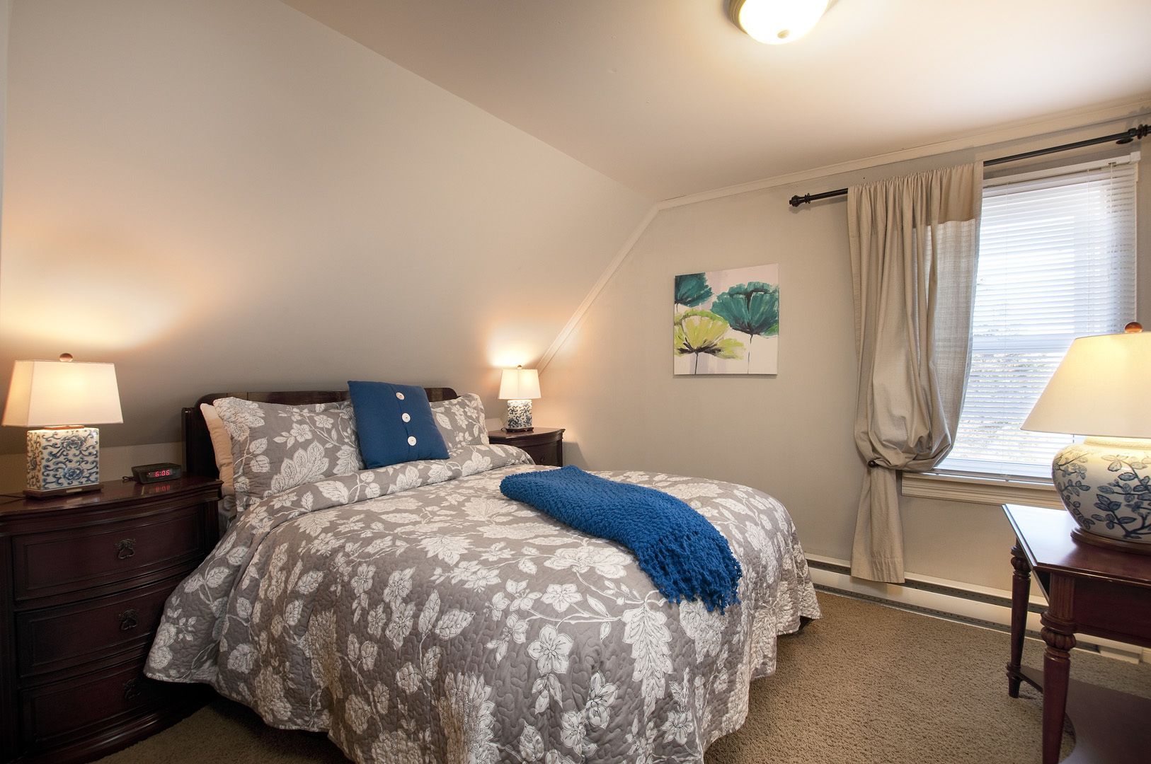 Un dormitorio con cama, mesitas de noche, lámpara, un cuadro y una ventana con cortinas.