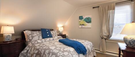Un dormitorio con cama, mesitas de noche, lámpara, un cuadro y una ventana con cortinas.