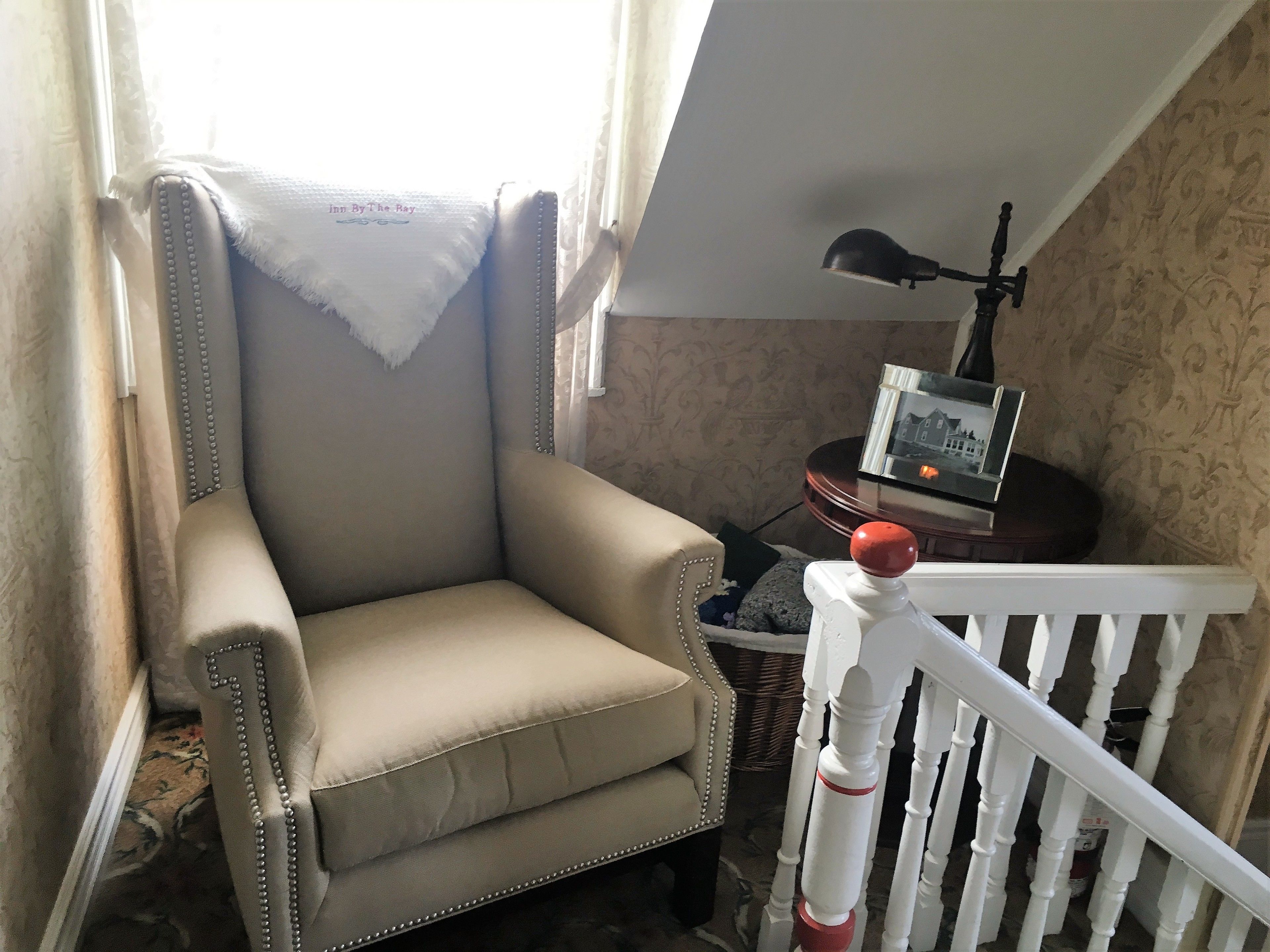 A corner room with a beige armchair, a white railing, a lamp, and a framed picture on a table.