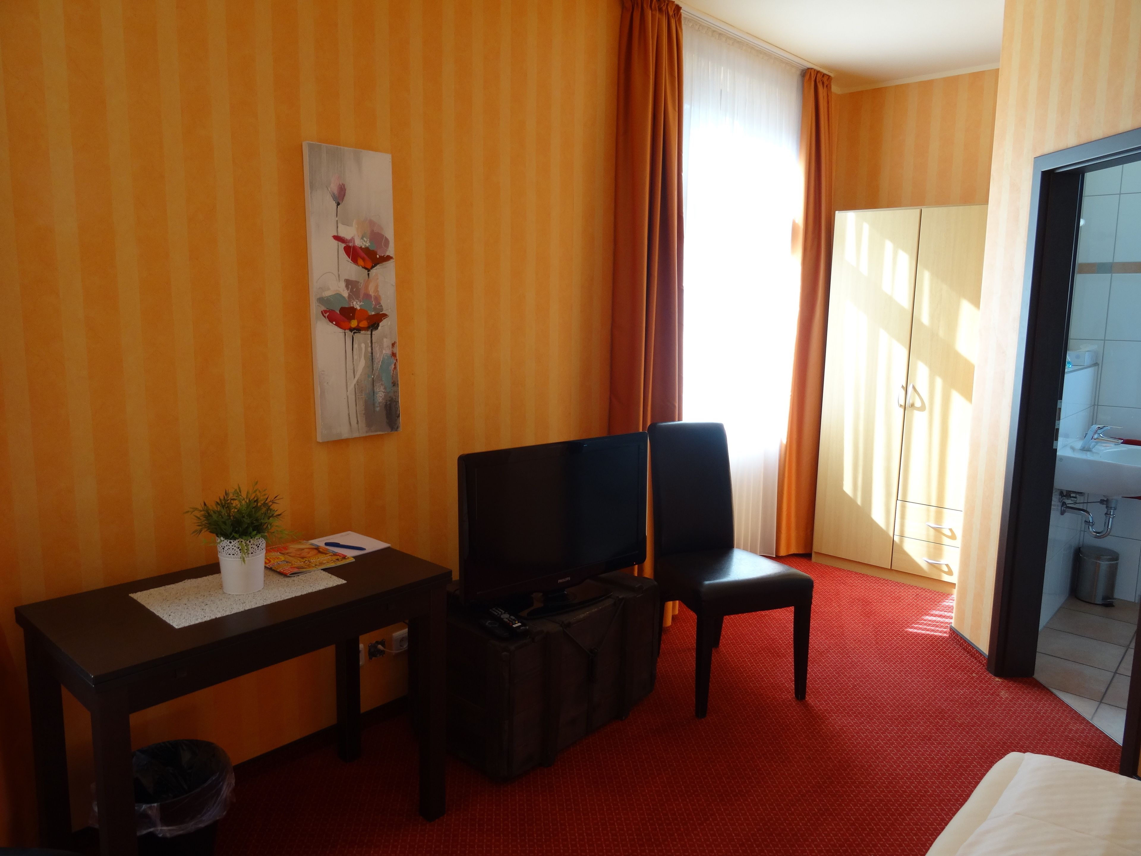 A hotel room with a bed, a desk with a television, a small plant, and a painting on the wall.