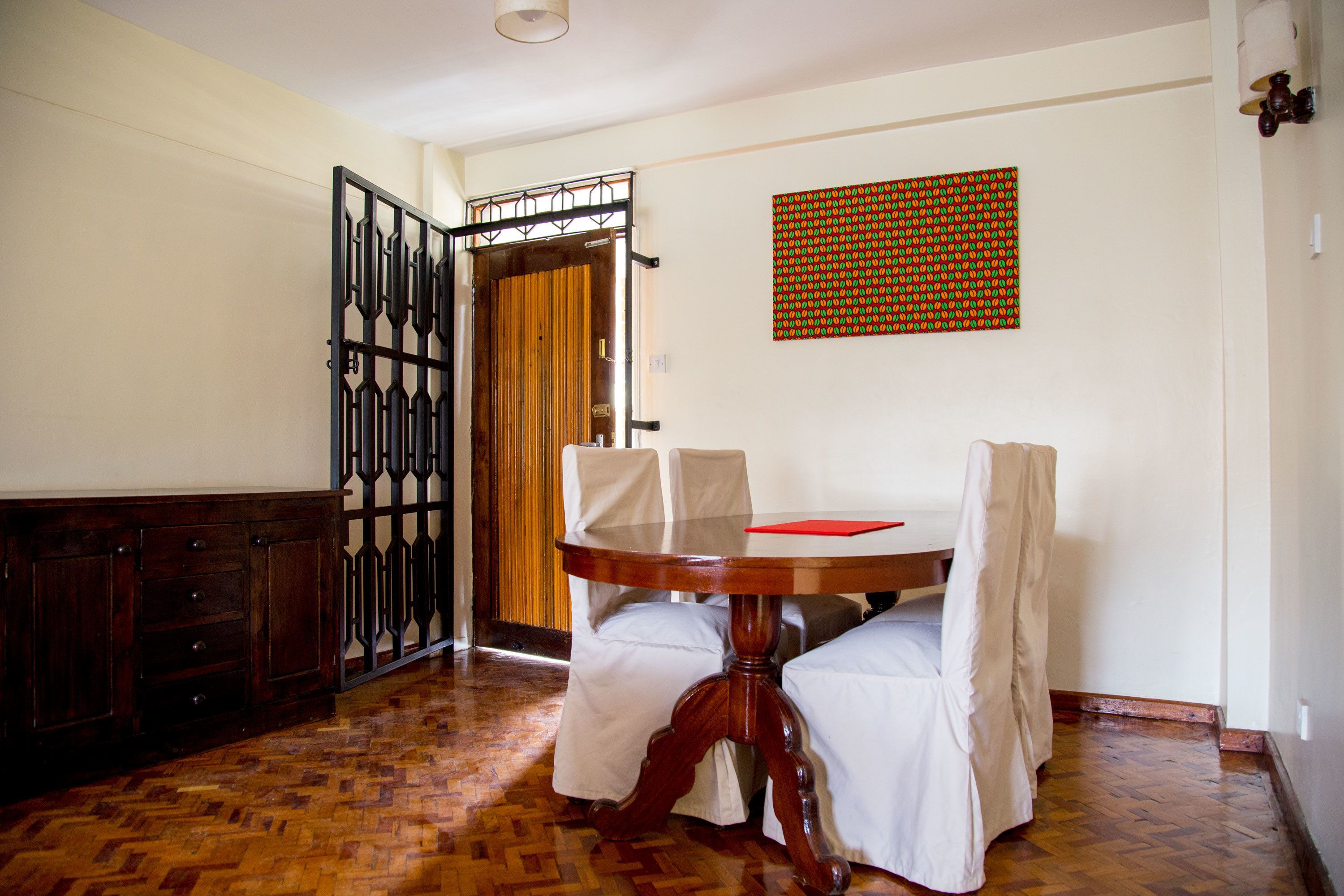 A room with a wooden floor, a round table with chairs, a door with a metal gate, and a patterned wall hanging.