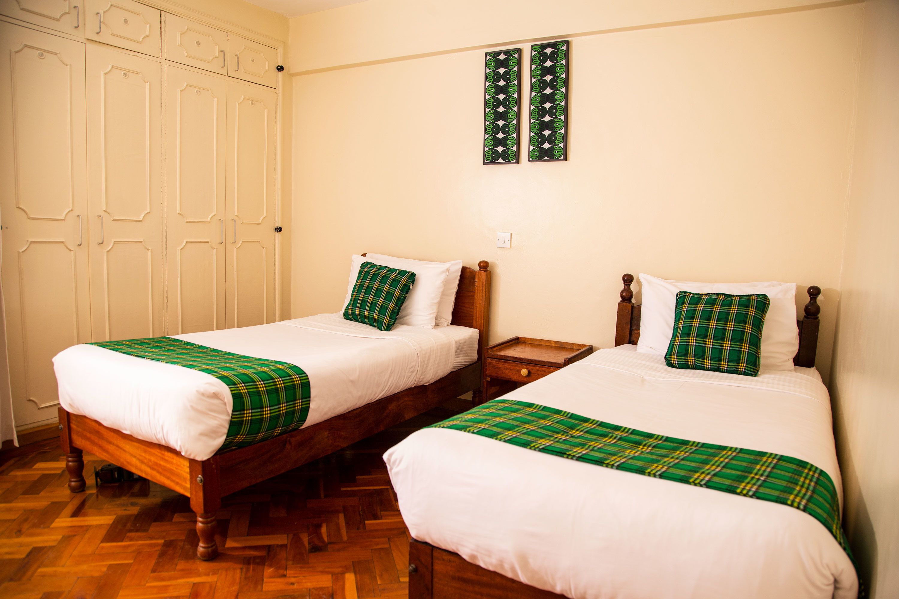 Two single beds with wooden frames, white bedding, and green plaid blankets. There are wooden nightstands with drawers, a white wardrobe, and two wall-mounted picture frames.