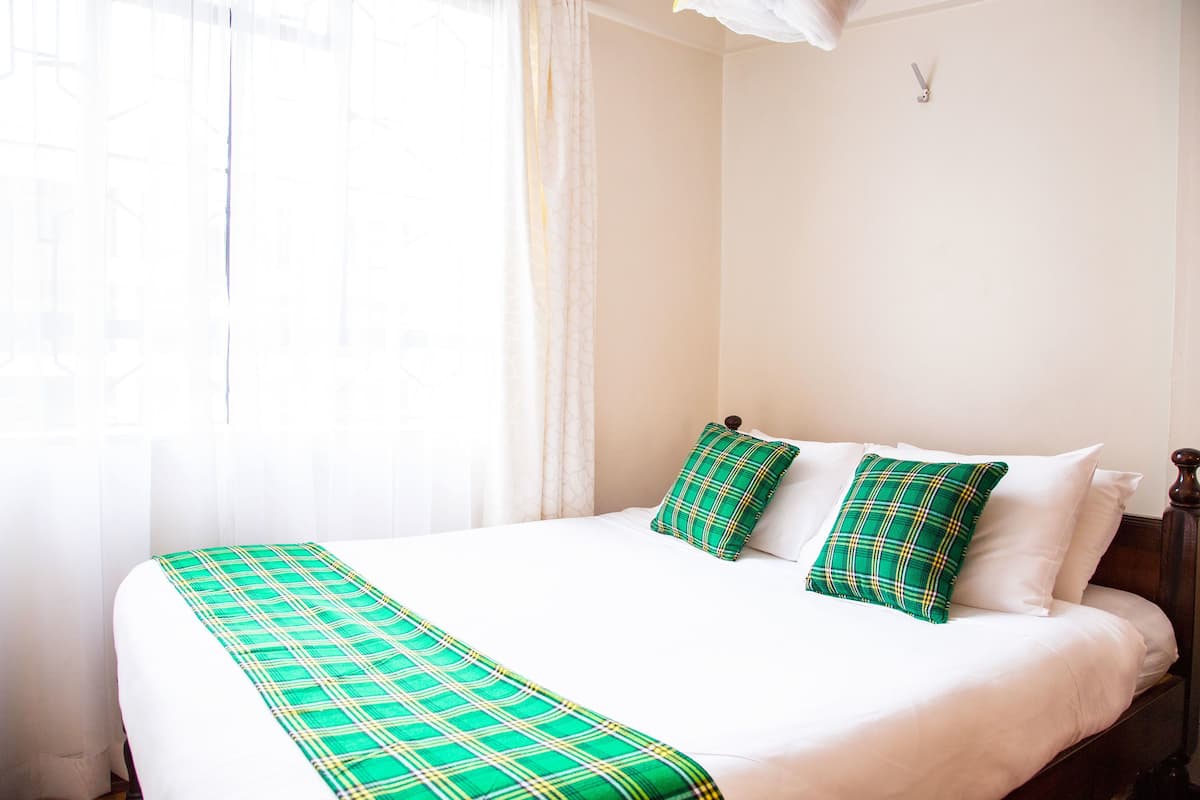 A neatly made bed with a green plaid bedspread and pillows, a wooden headboard, and a window with sheer curtains.