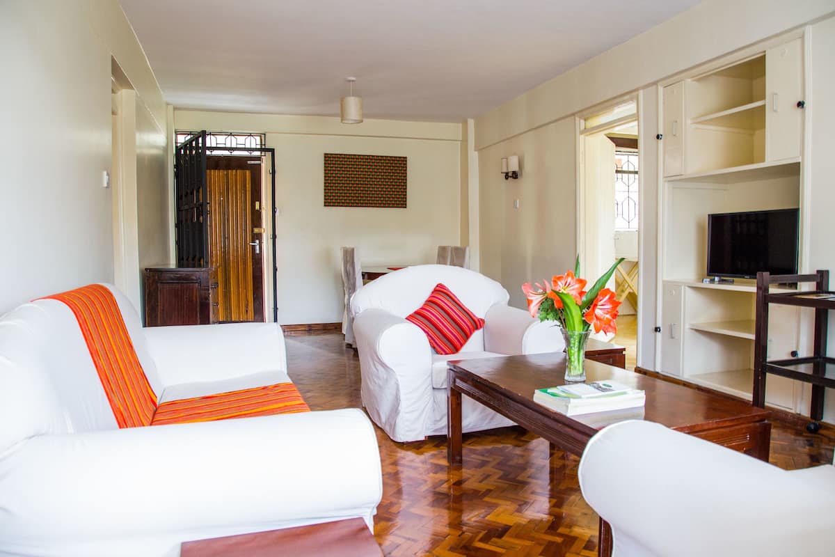 A hotel room with a white sofa, a red pillow, a wooden coffee table, and a TV on a stand.