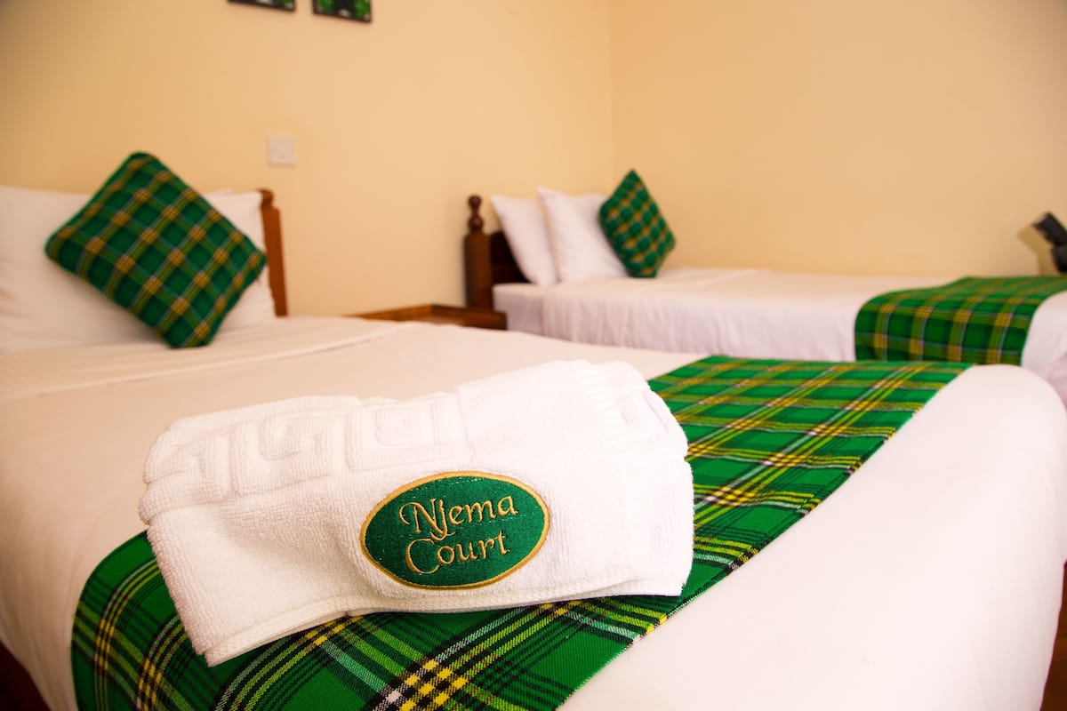 A hotel room with two beds, each with a green plaid bedspread and matching pillows. A white towel with "Nyema Court" embroidered on it is placed on the bed in the foreground.