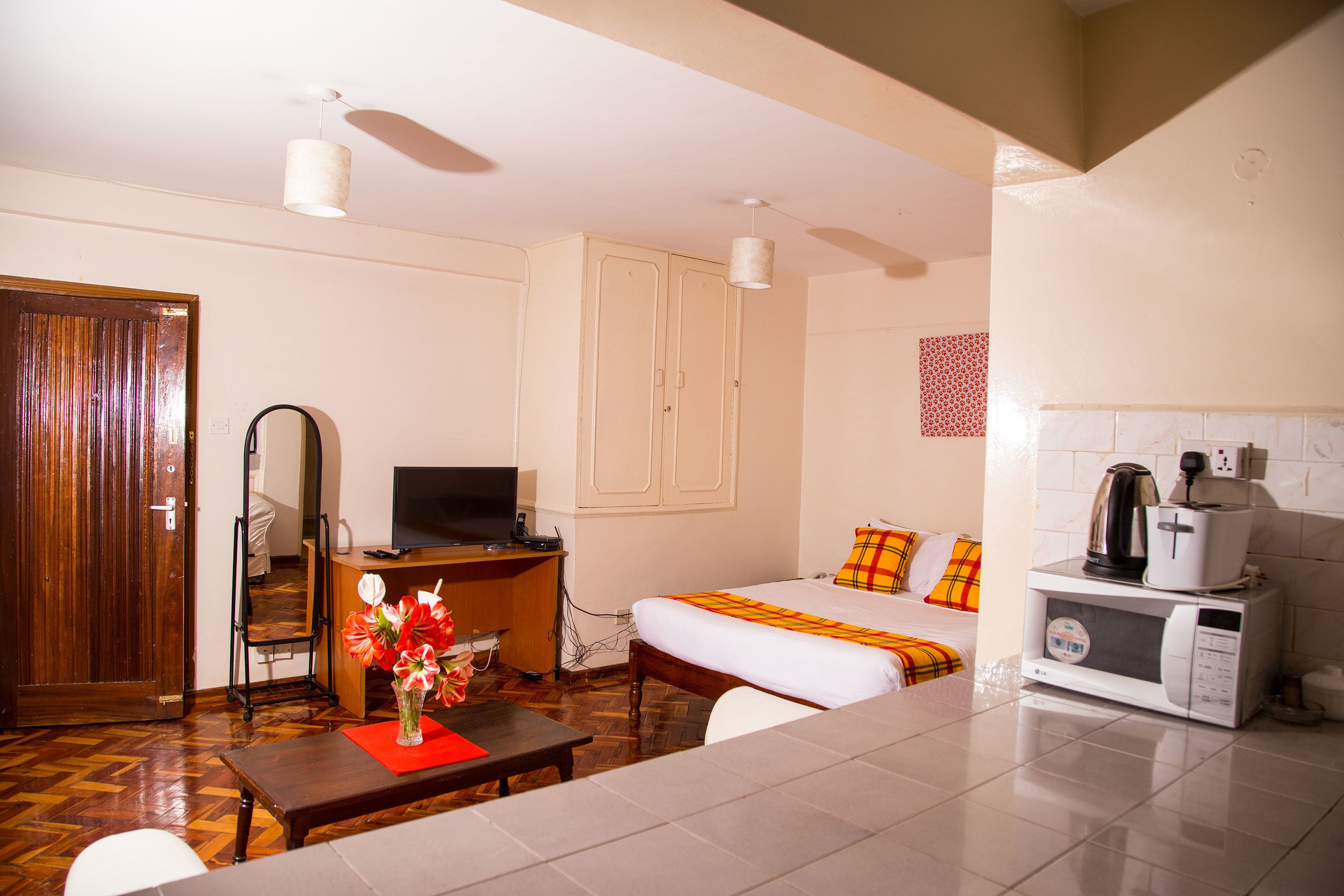 Habitación de hotel con escritorio de madera, televisor, cama con manta de cuadros, microondas y pequeña mesa con un jarrón de flores.