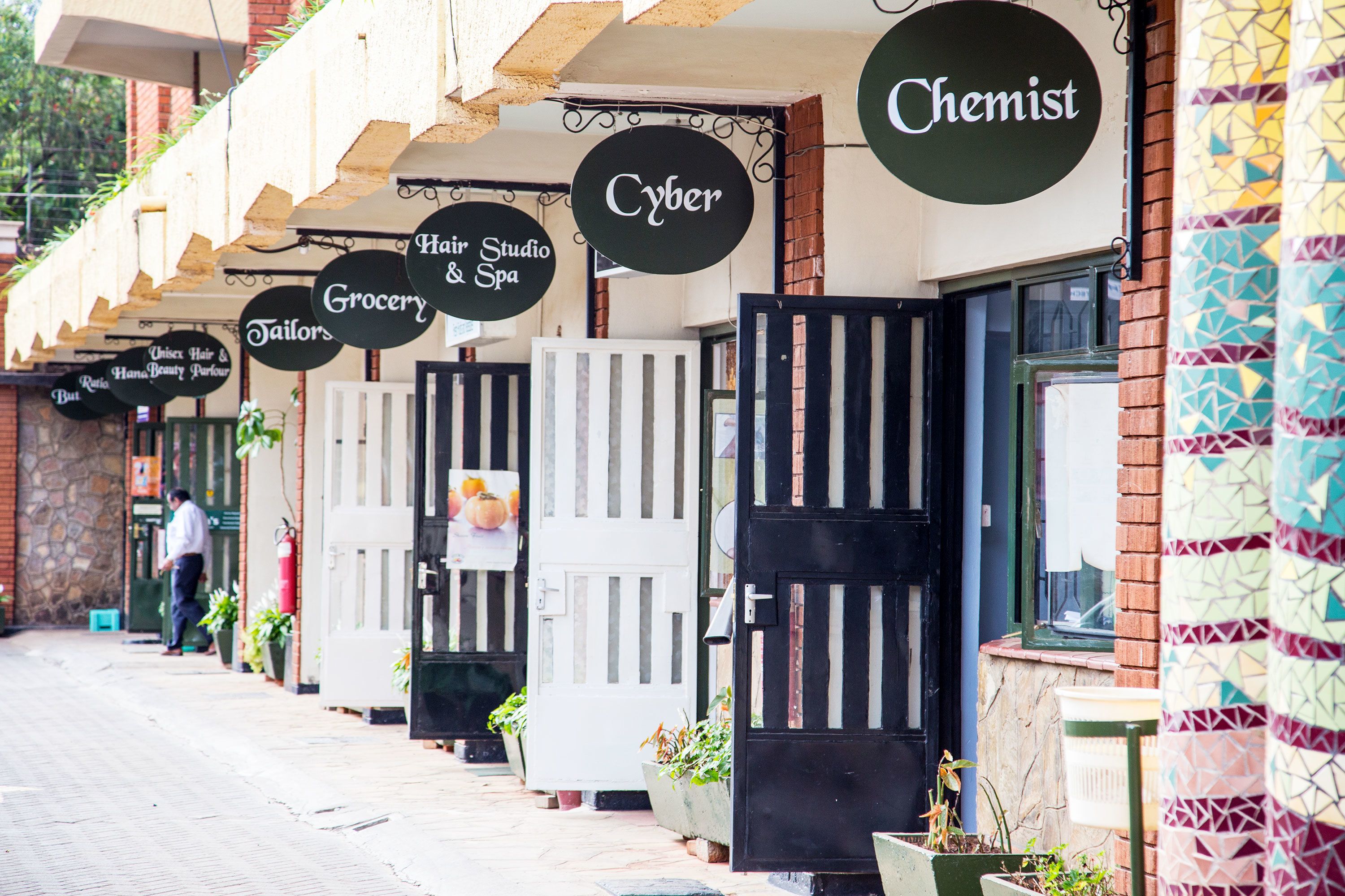 A row of storefronts with signs for 'Hair Studio & Spa', 'Grocery', 'Tailors', and 'Chemist'.
