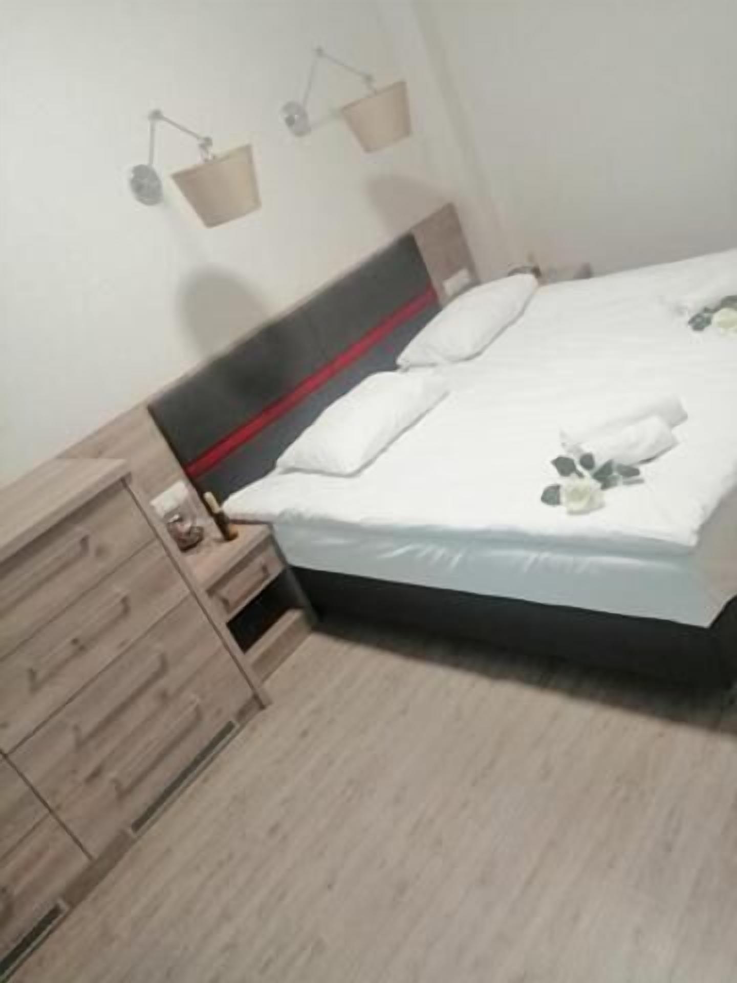 Un lit bien fait, avec une couette blanche et des oreillers, une table de chevet en bois avec une lampe, et une commode en bois avec des tiroirs.