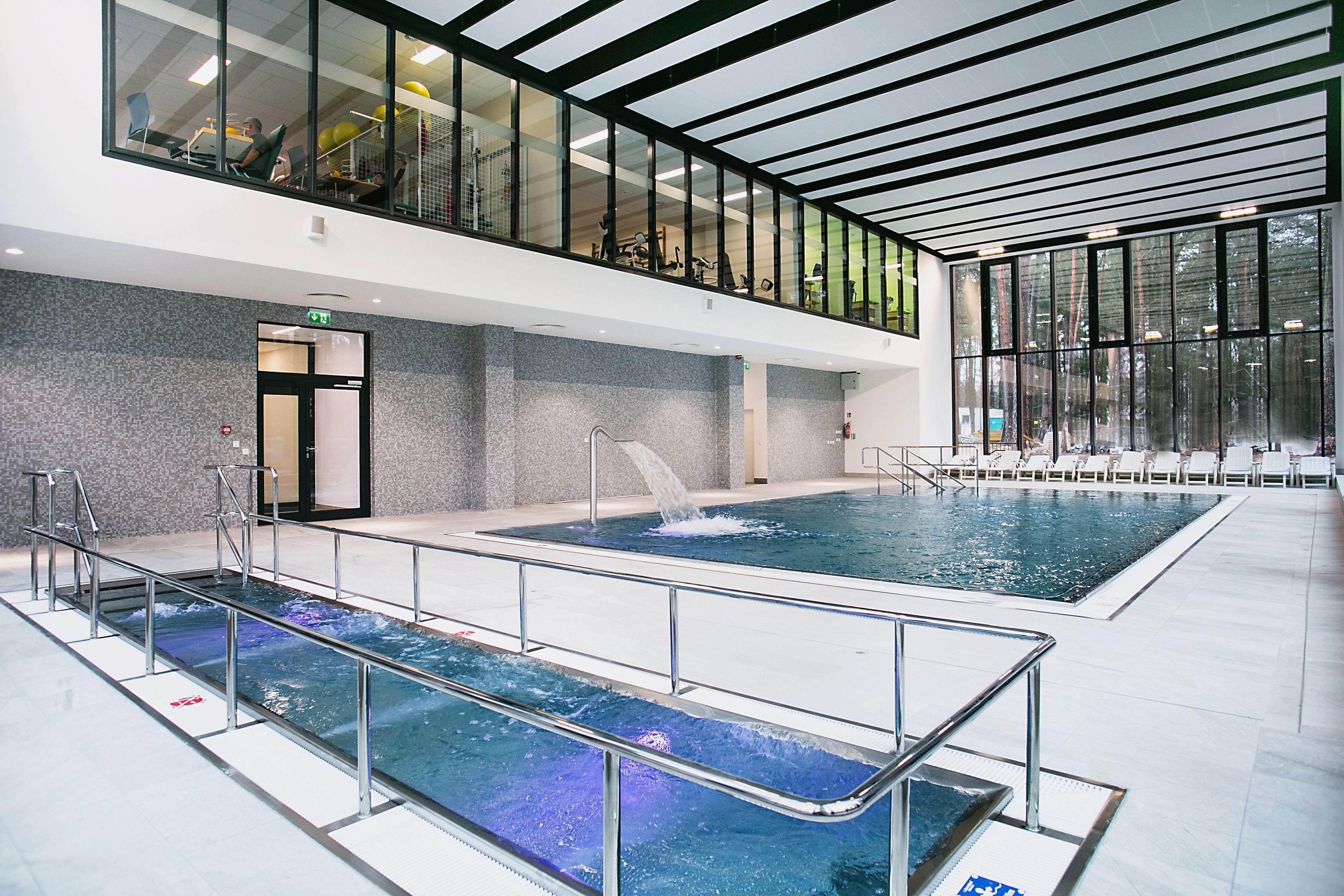 A modern indoor swimming pool with multiple lanes, a viewing area above, and a sunlit space with seating.