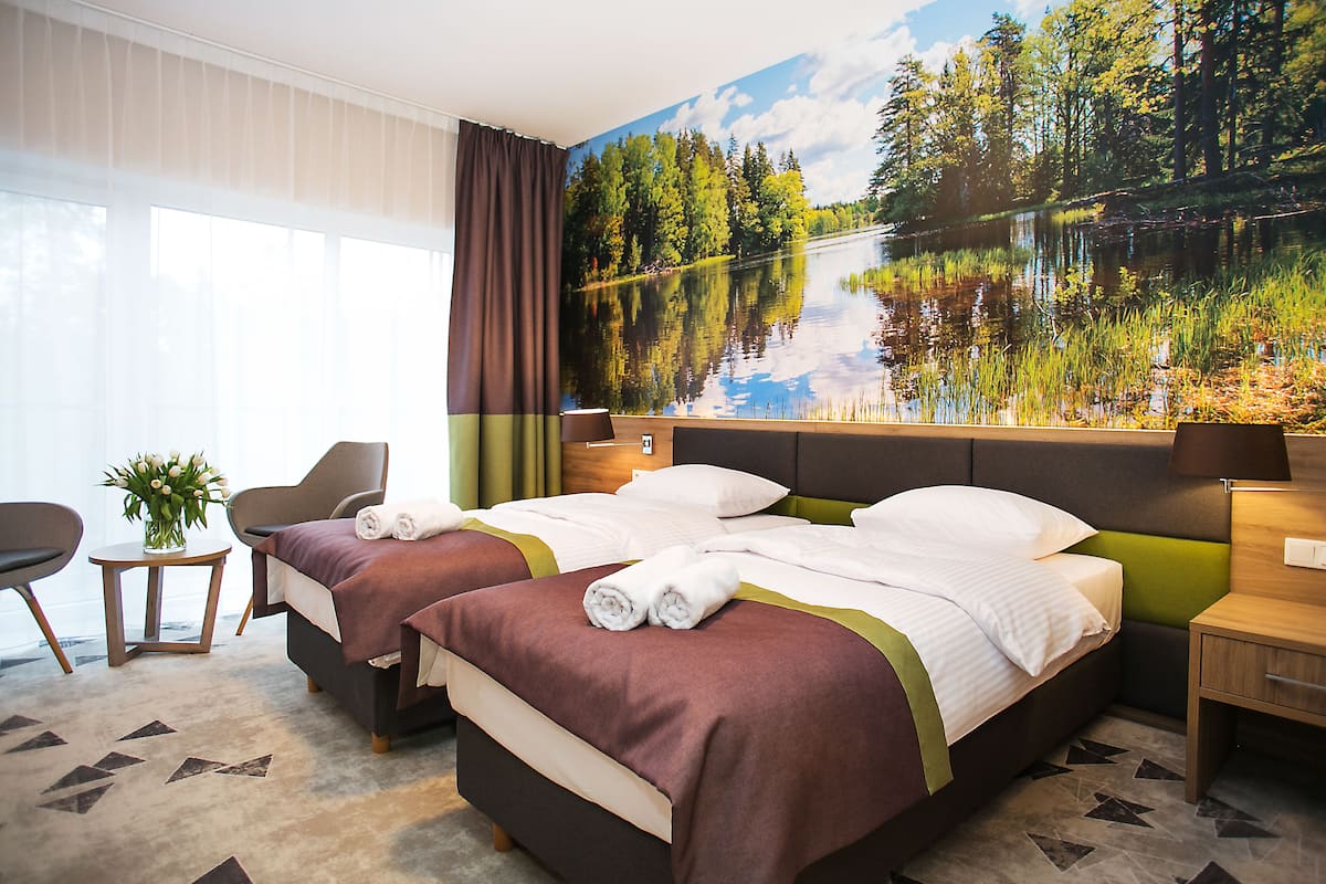 Habitación de hotel con dos camas, un ventanal grande, un cuadro de un lago y una mesita con un florero.