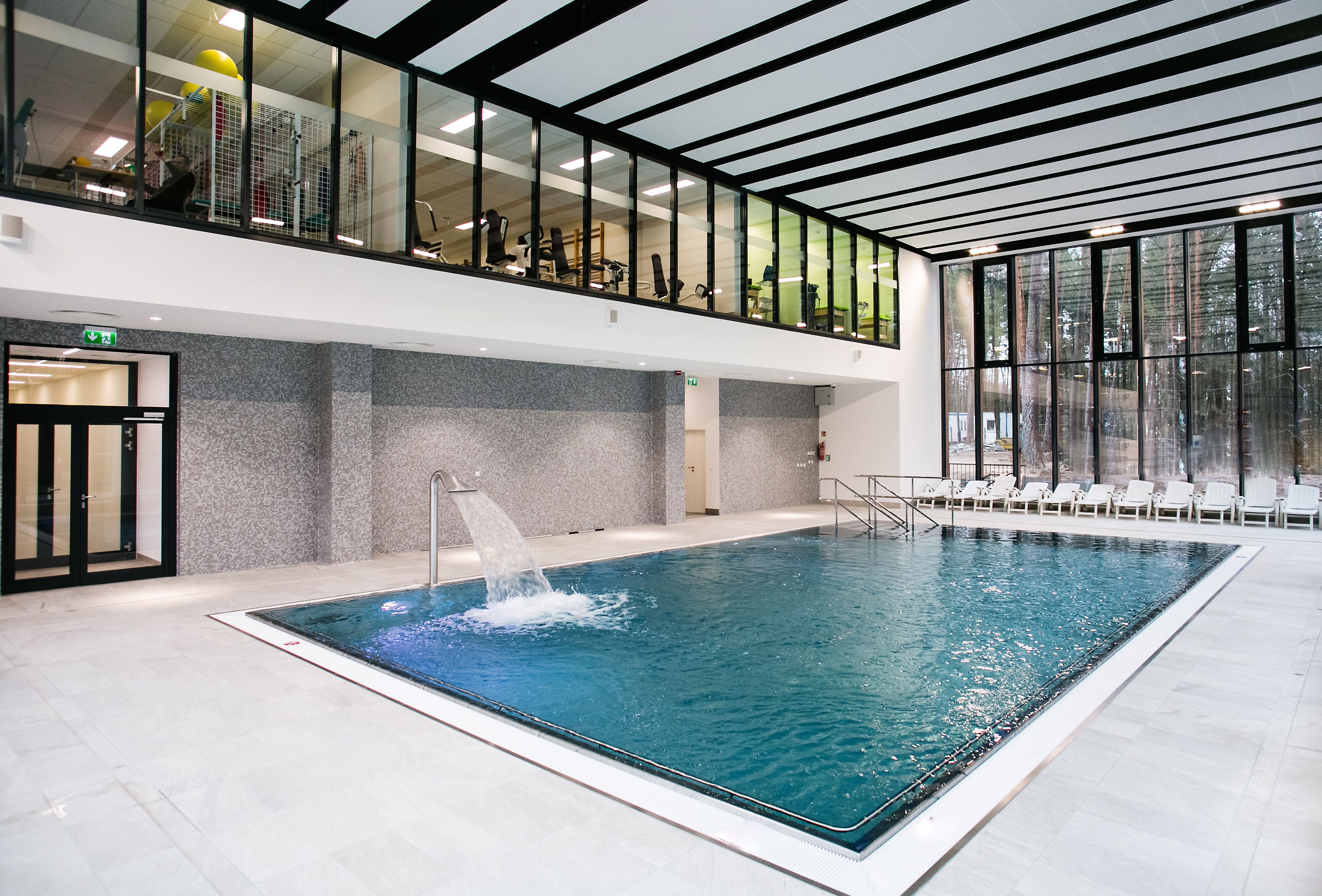 A large indoor swimming pool with a water feature, a glass balcony, and a seating area with chairs.