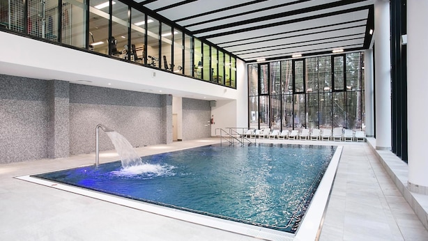Una piscina interior moderna con techo de vidrio, una fuente de agua y un área de descanso con sillas.