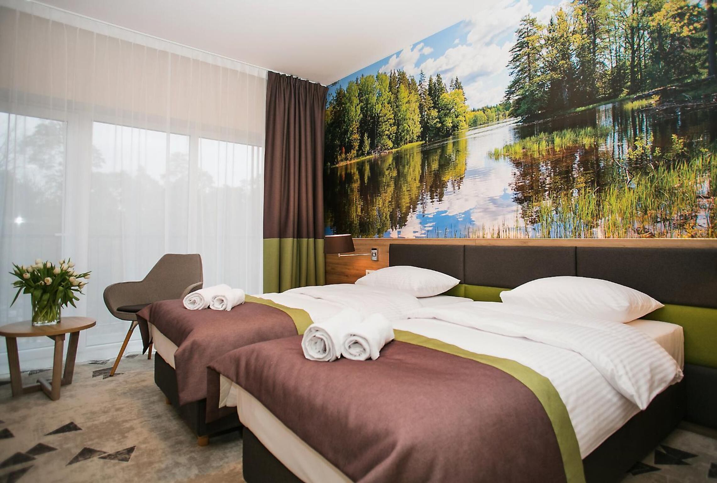 A hotel room with two beds, a large window with sheer curtains, a painting of a lake, and a small table with a vase of flowers.