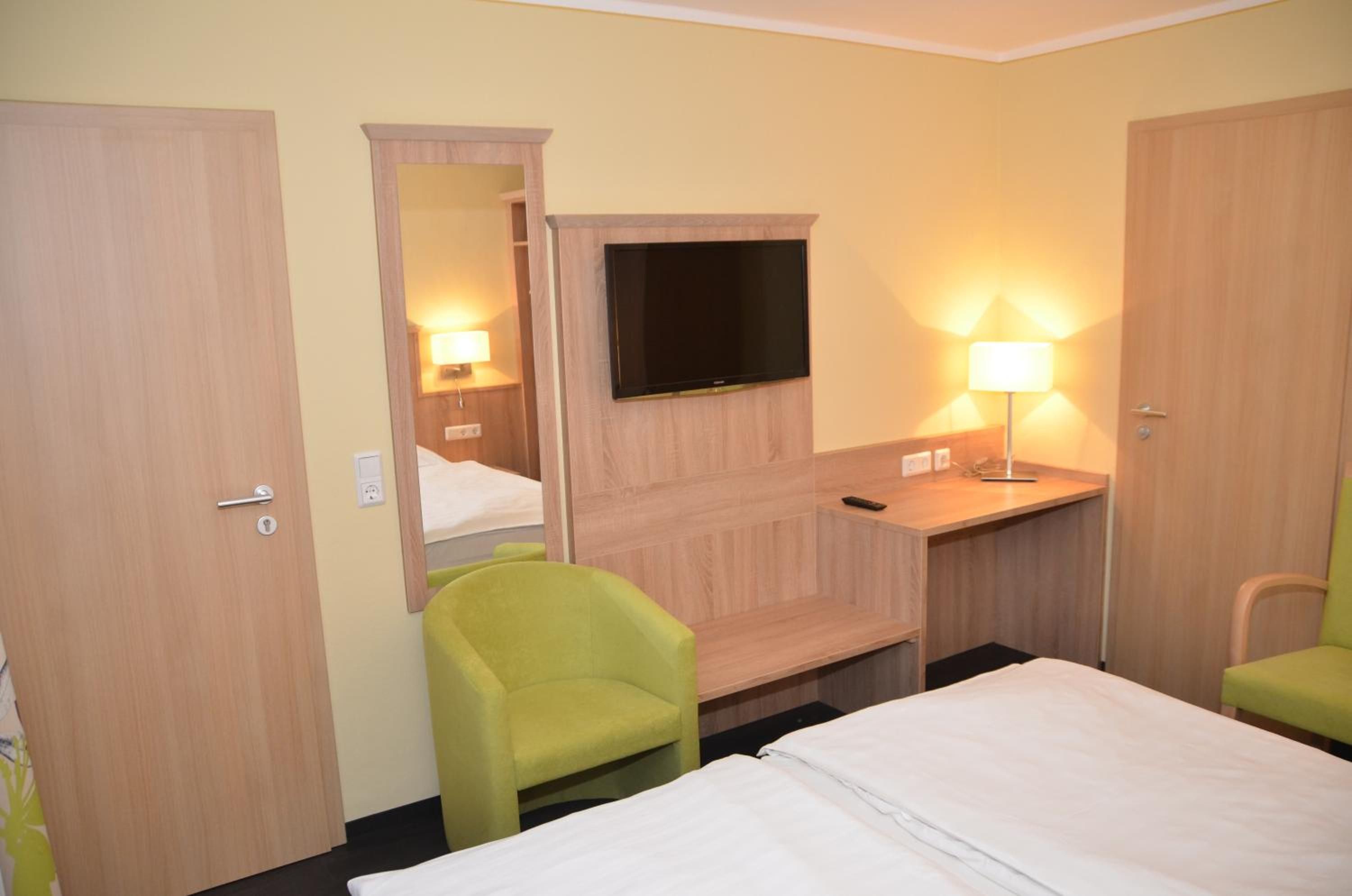 A hotel room with a bed, a desk with a flat-screen TV, two chairs, and a mirror.