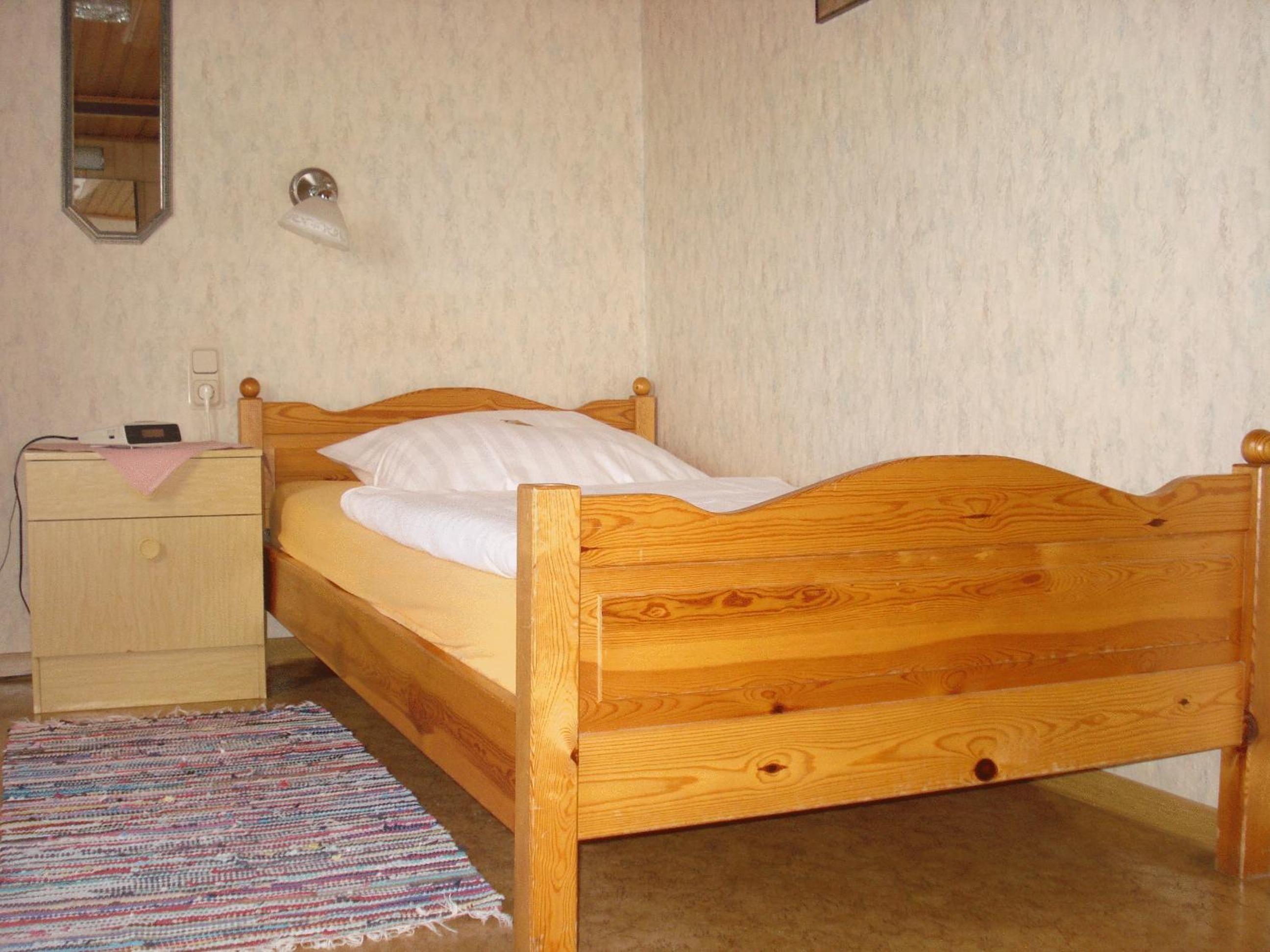 A single bed with a wooden frame, a bedside table, a small rug, and a wall-mounted light fixture.