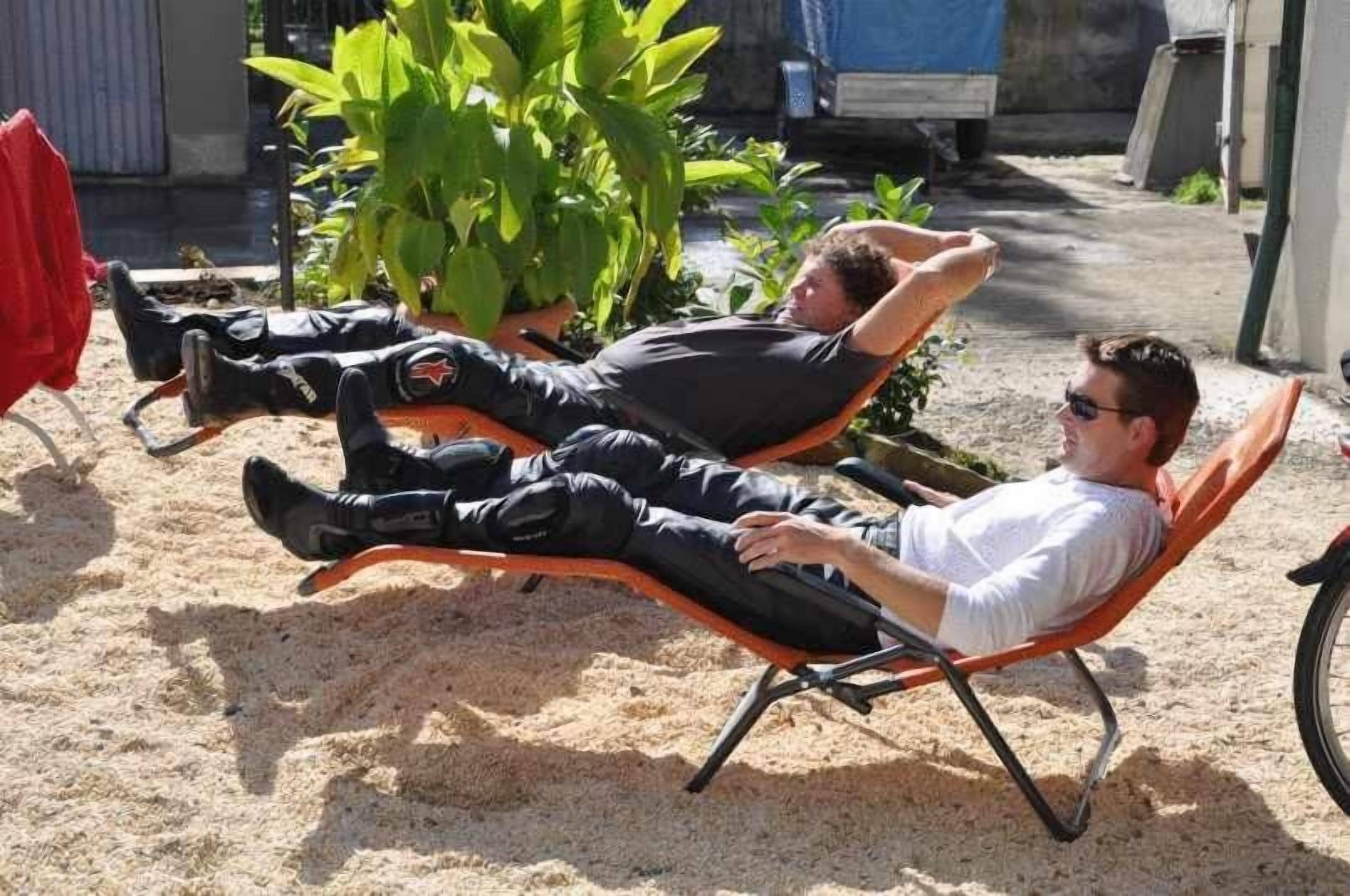 Two individuals reclining on lounge chairs outdoors with a plant and a red object nearby.