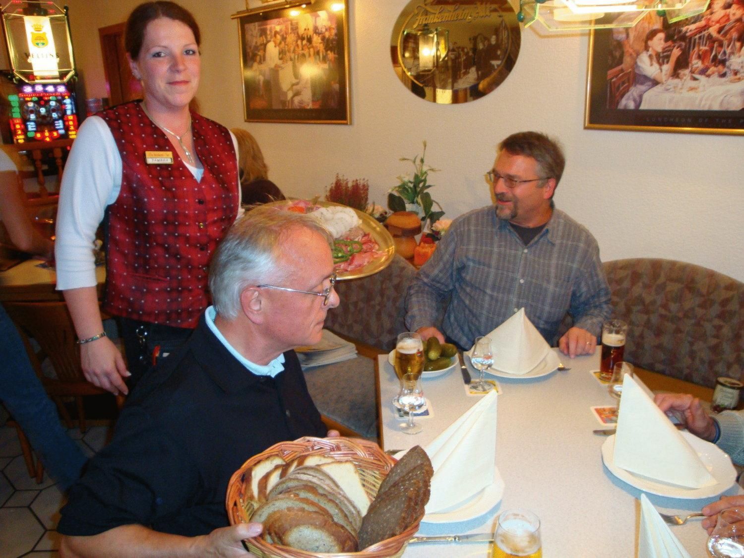 A dining area with a table set for four, including a basket of bread, plates, and glasses of beer.