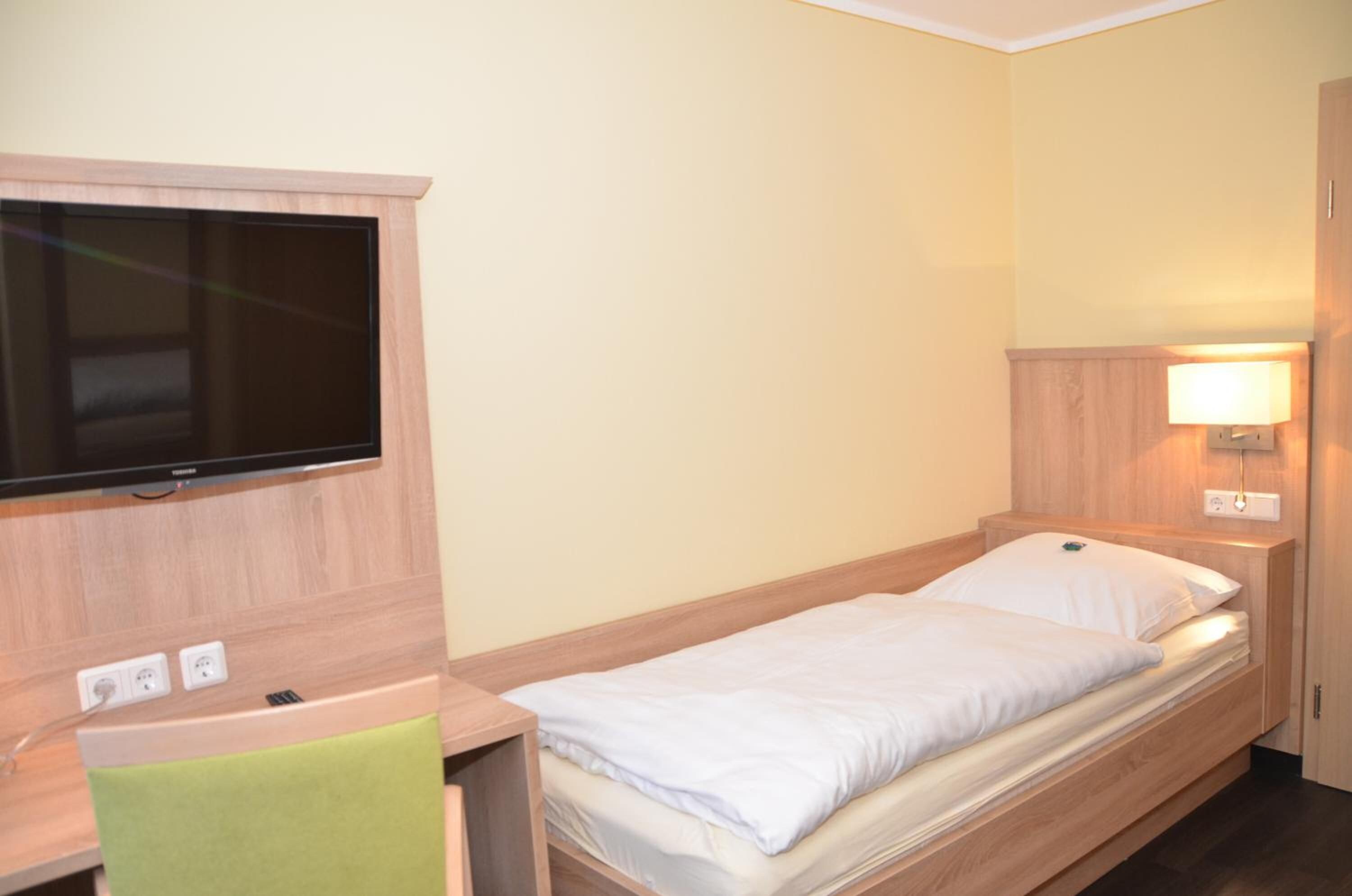 A single bed with a wooden headboard, a bedside table with a lamp, and a flat-screen TV mounted on the wall.