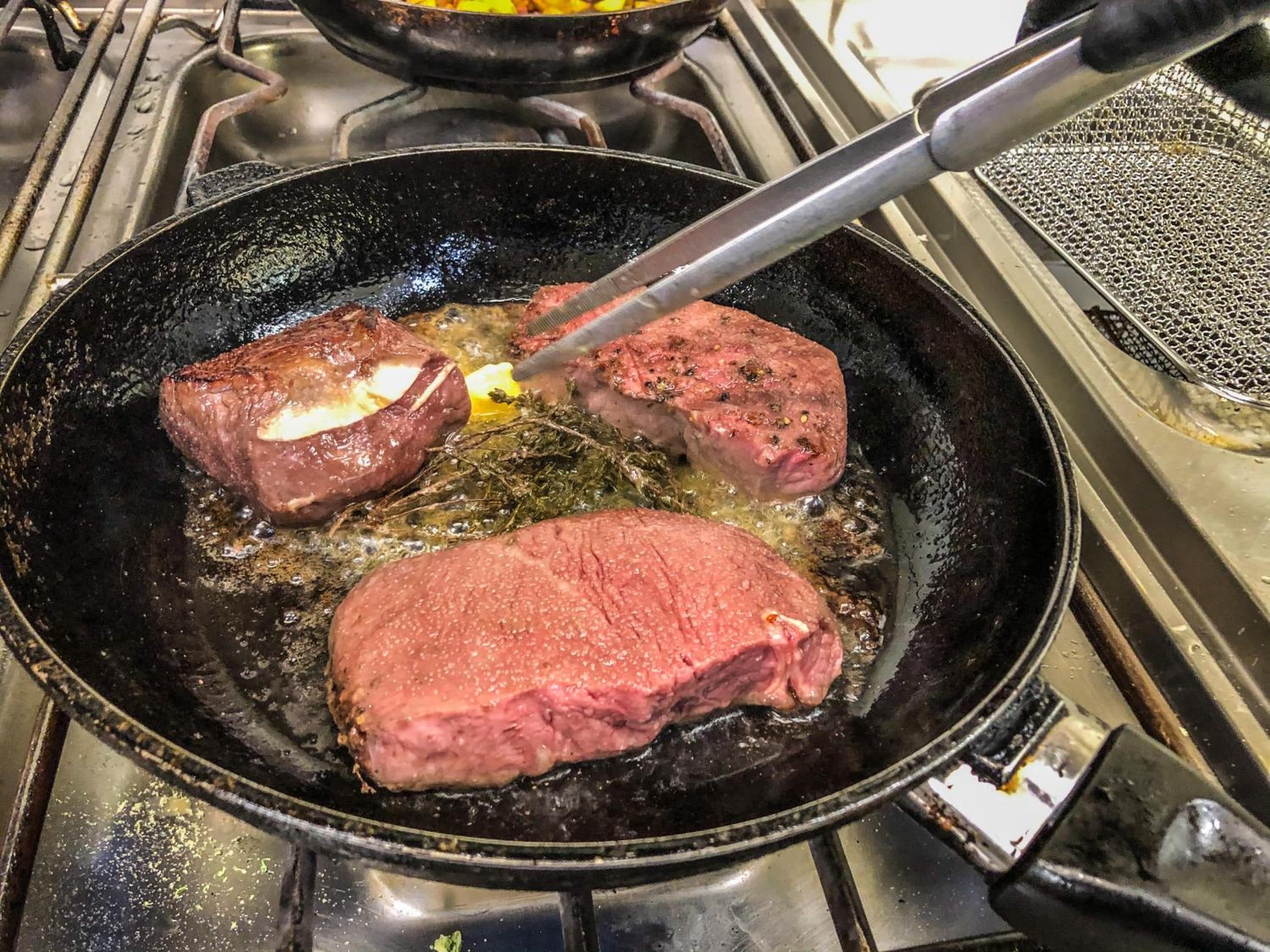 En stekpanna står på spisen och det ligger två bitar kött i som steks.