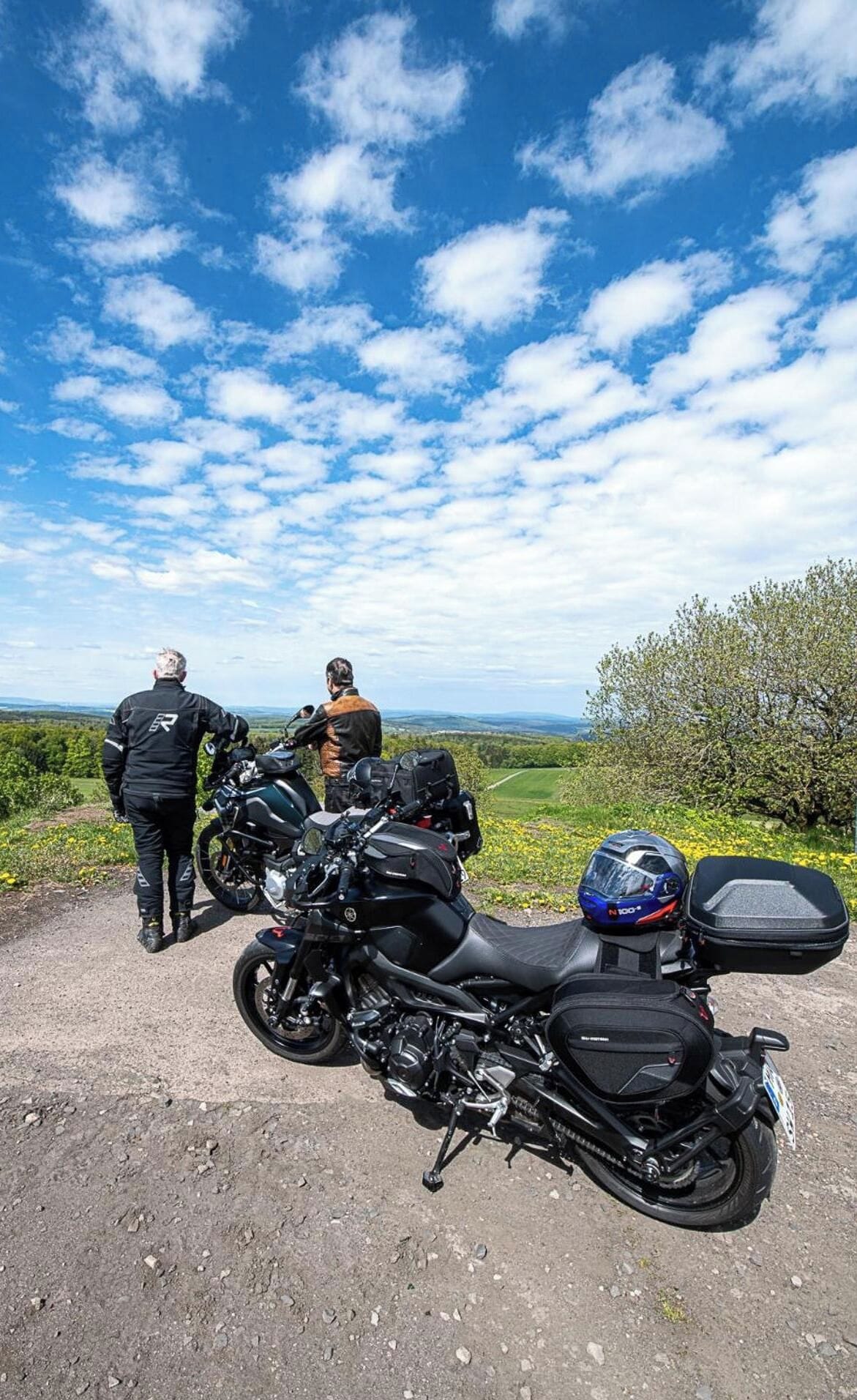 Two motorcycles parked on a dirt path with a scenic view of green fields and trees under a blue sky with clouds.