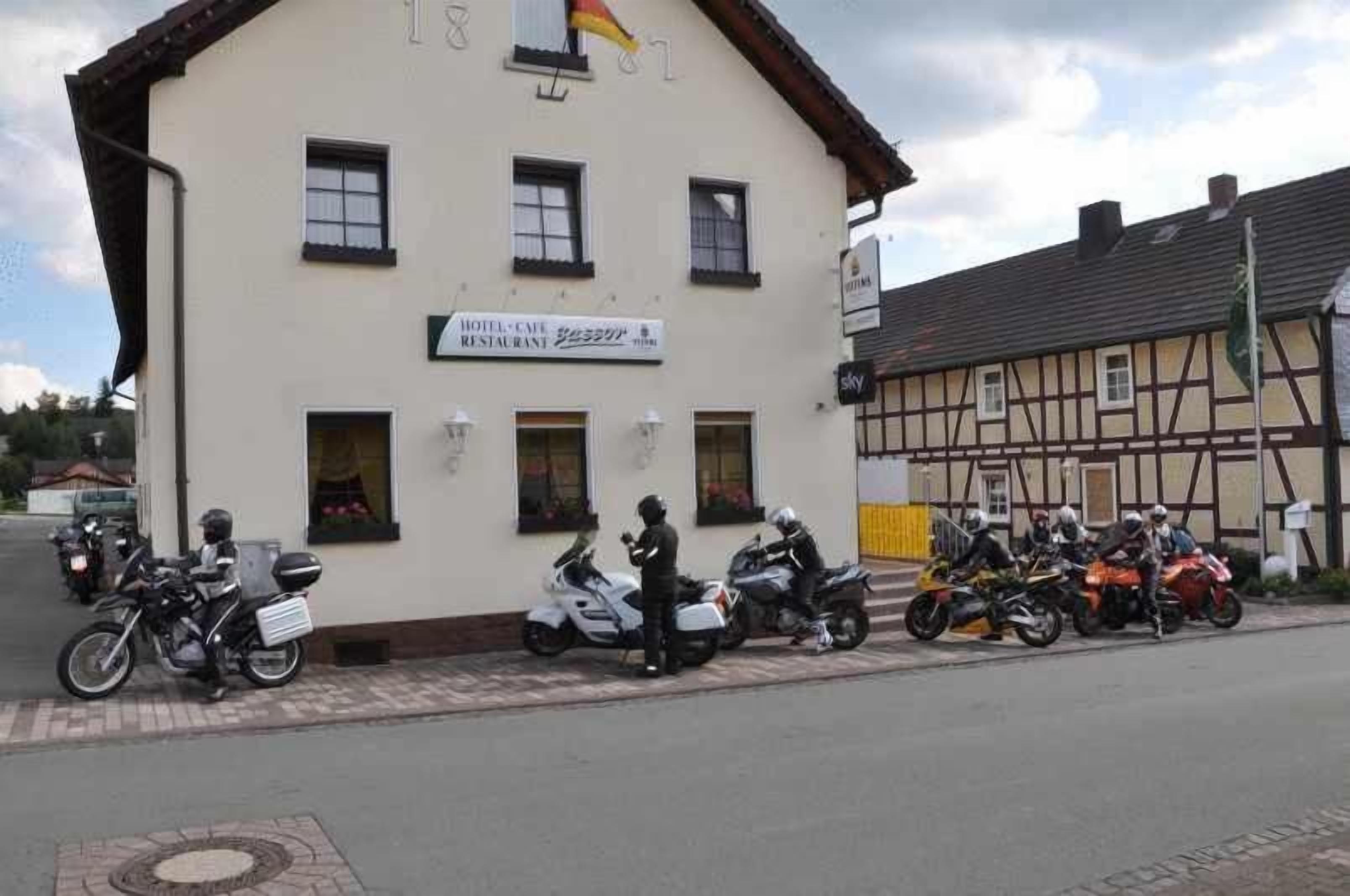 Un bâtiment sur lequel on peut voir une enseigne indiquant qu'il s'agit d'un hôtel et d'un restaurant, plusieurs motos garées devant, et une structure traditionnelle à colombages en arrière-plan.