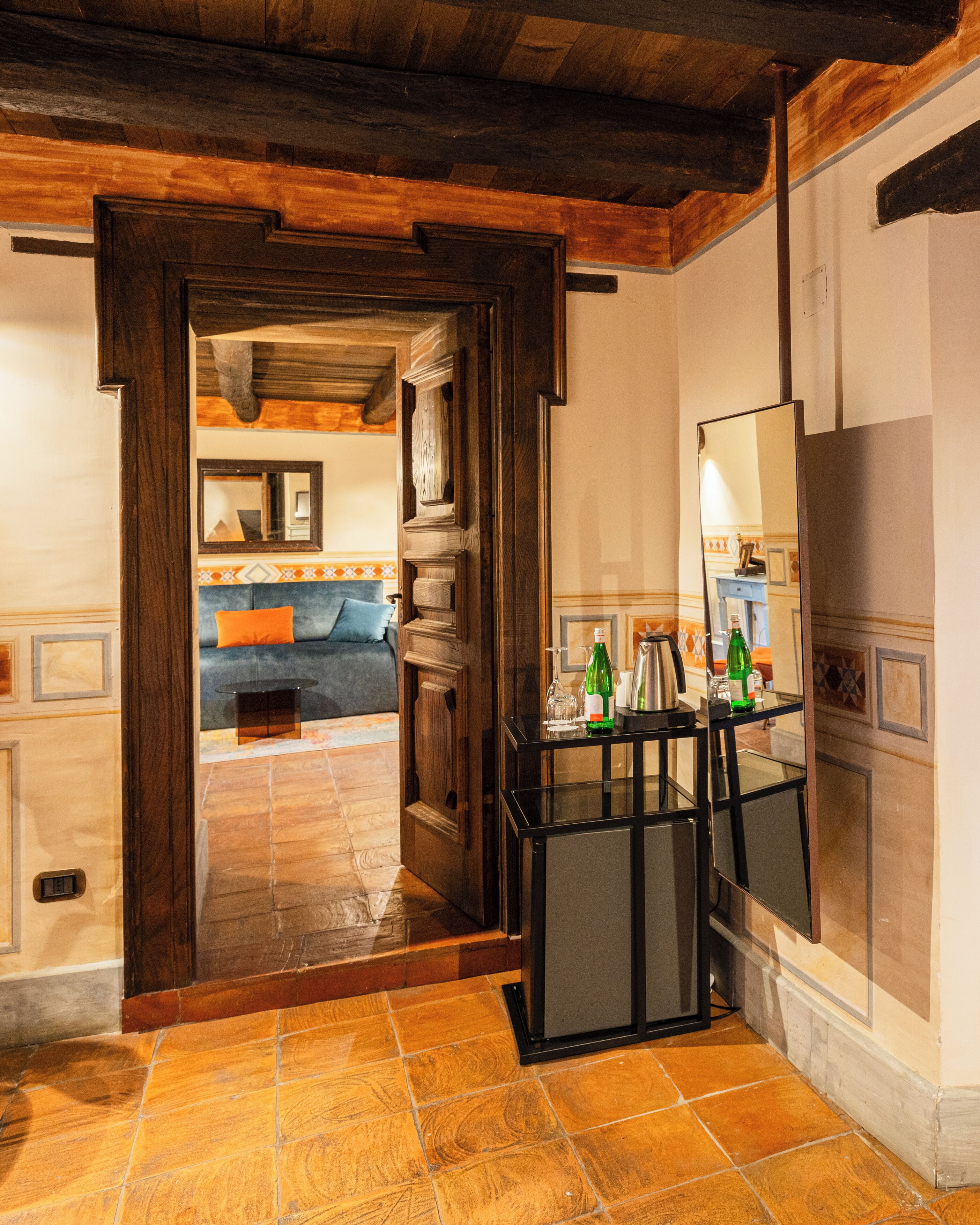 Une pièce avec un plafond en bois, un sol carrelé, un lit avec des coussins bleus et orange, une petite table avec des bouteilles et un miroir au mur.