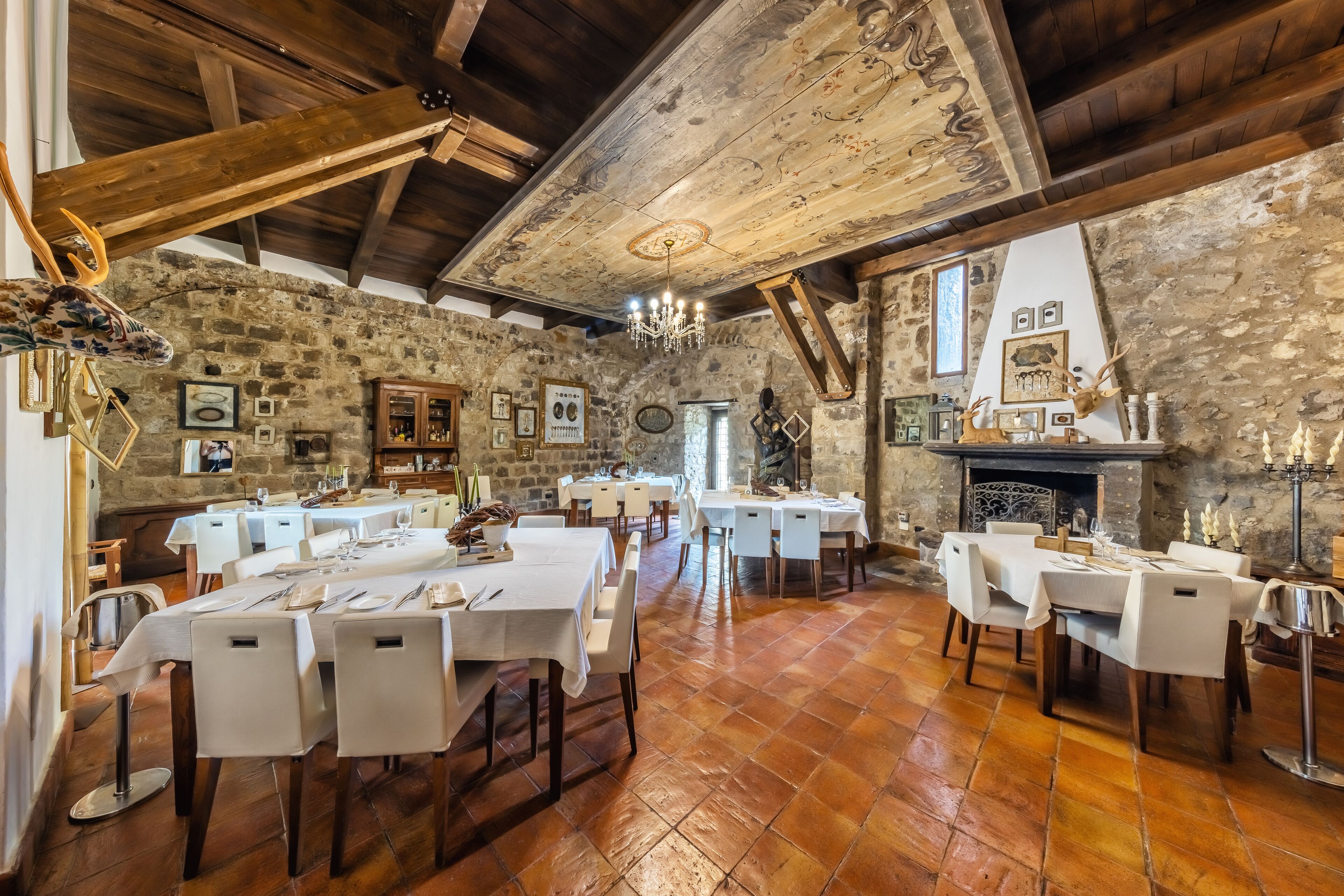 Un comedor rústico con paredes de piedra, vigas de madera y una chimenea. Las mesas están dispuestas con manteles blancos y sillas.