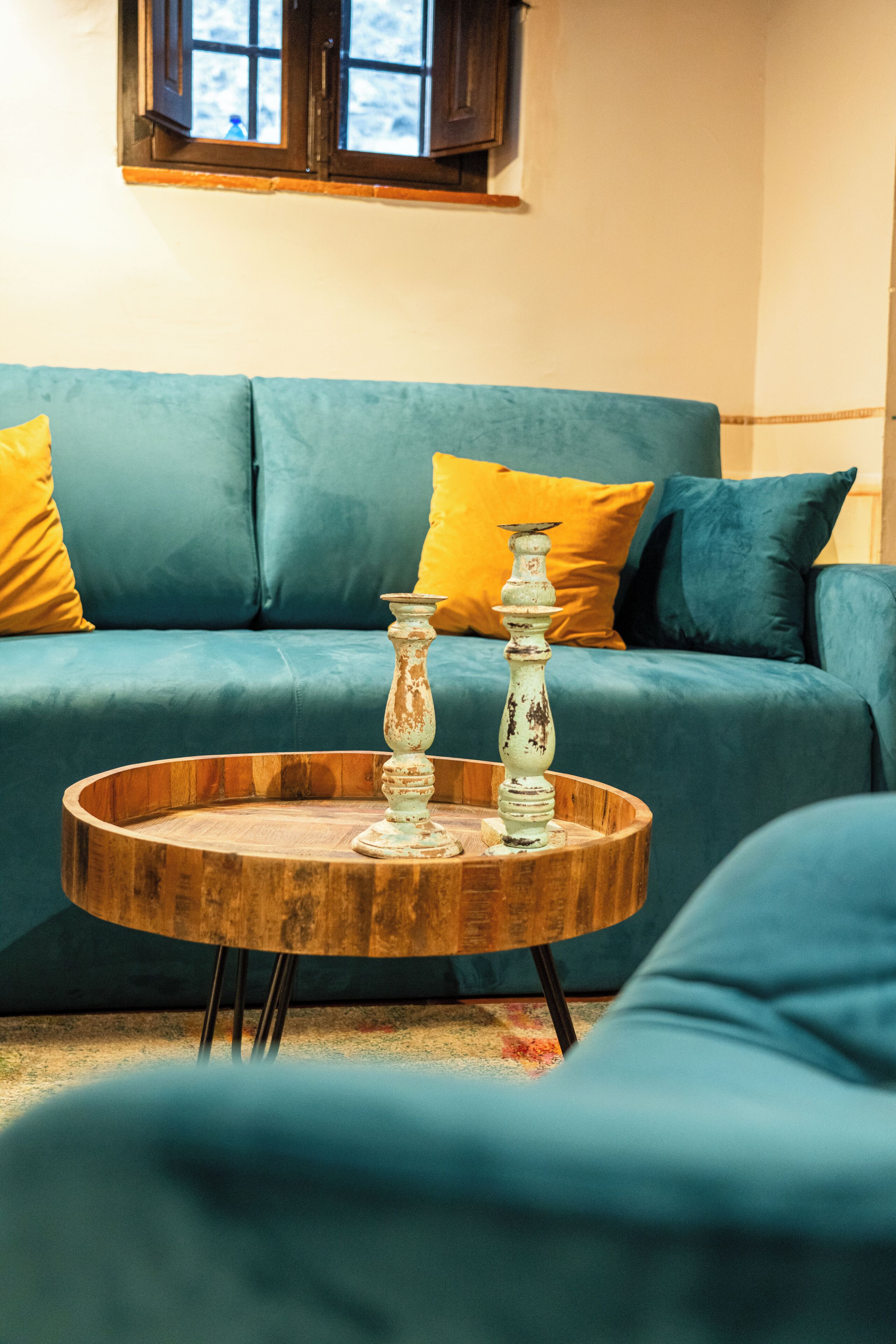 Un sofá color turquesa con cojines amarillos, una mesa de centro de madera y dos portavelas blancos, además de un ventanal.