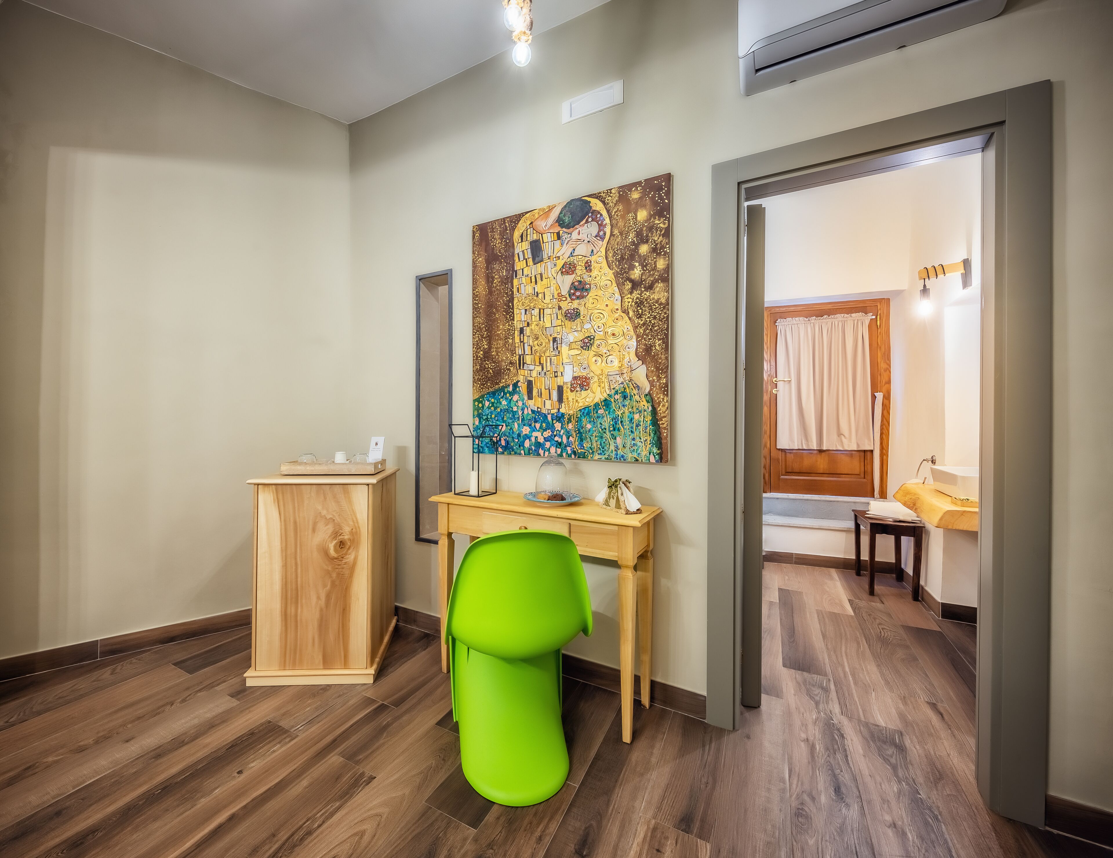 A hotel room with a wooden desk, a green chair, a painting on the wall, and a wooden wardrobe.