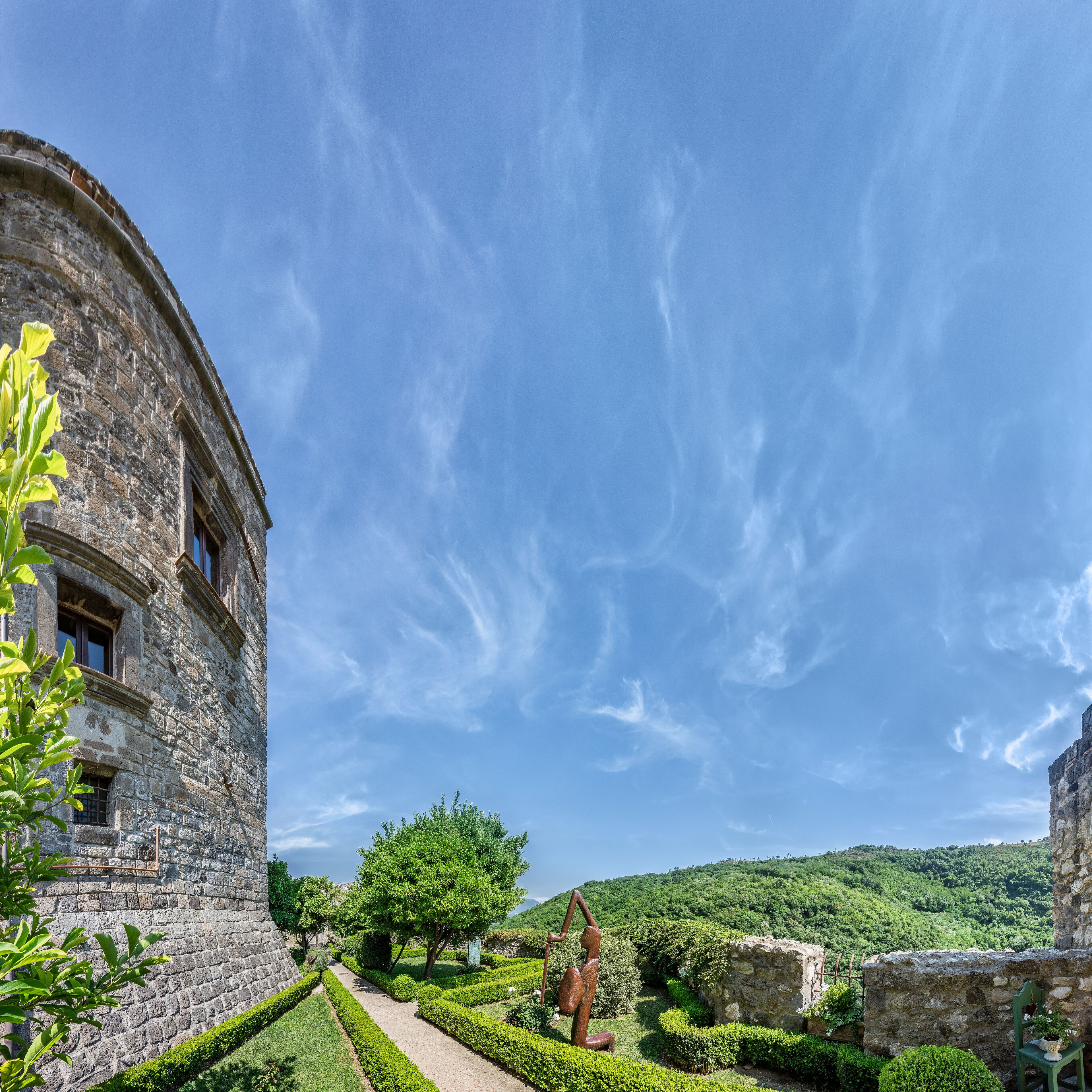 Un bâtiment historique en pierre, doté d’une tour circulaire, entouré de jardins bien entretenus et surplombé d’un ciel bleu azur.