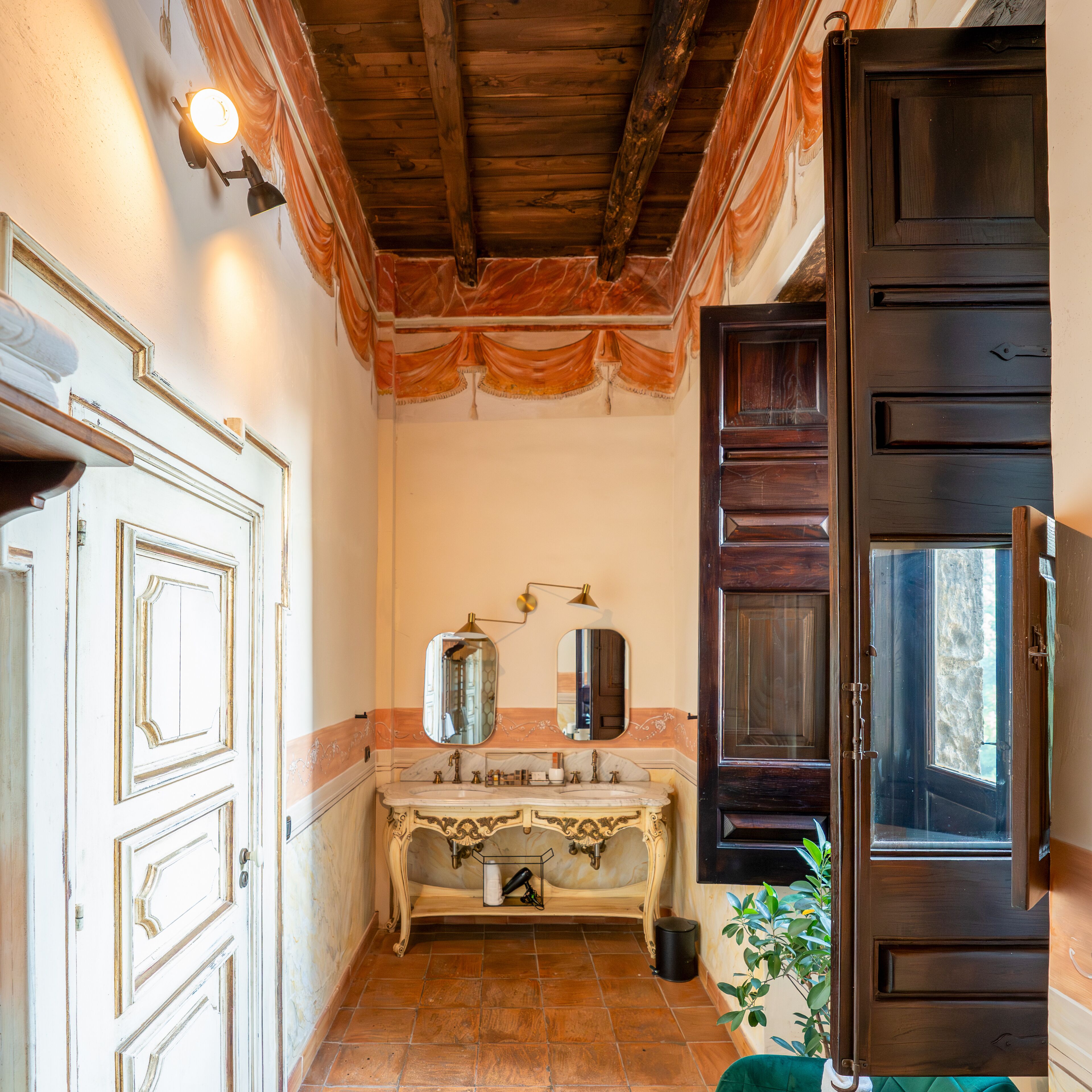 A narrow hallway with a vintage-style bathroom featuring a marble sink, ornate mirror, and a wooden door with a window.