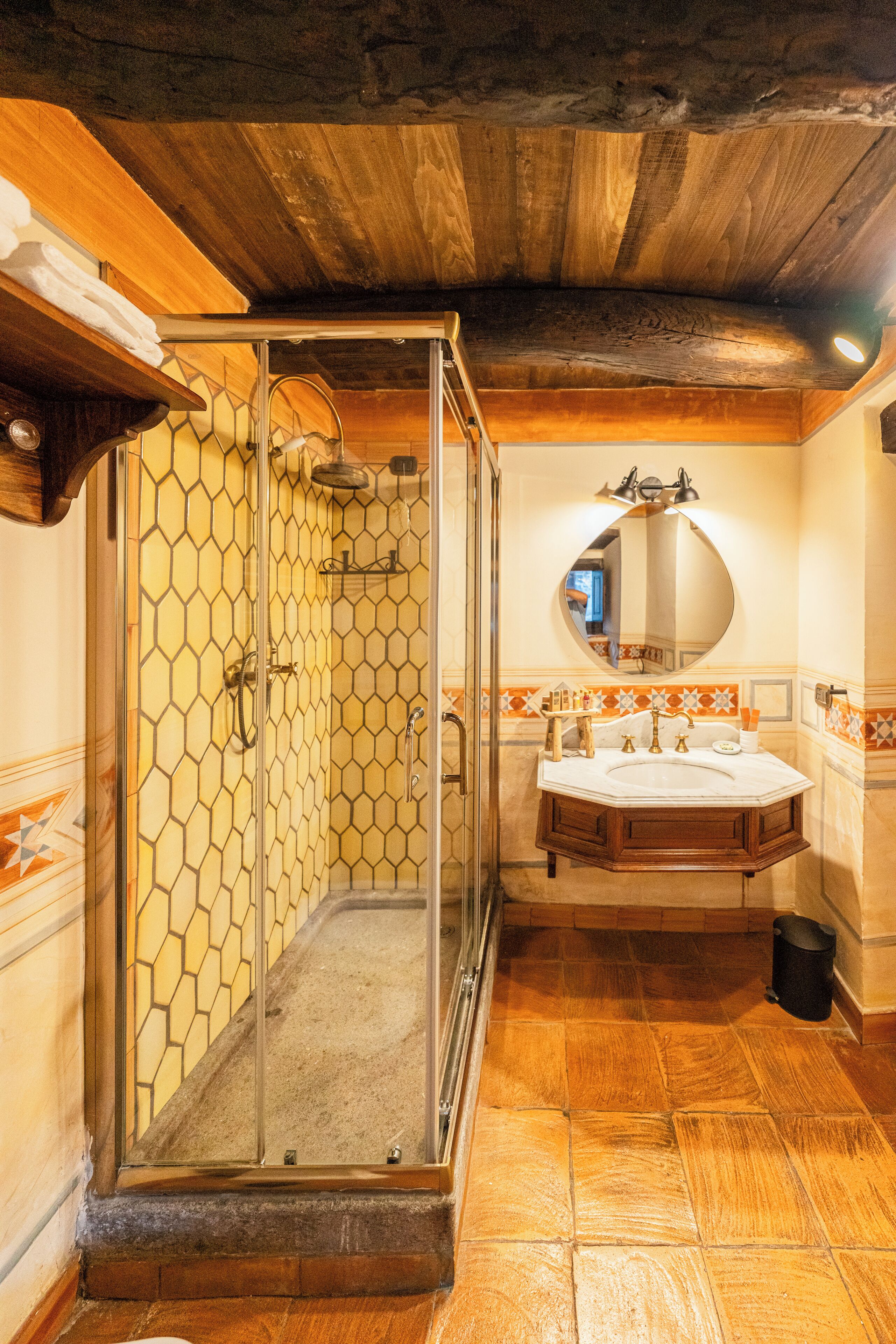Ein Badezimmer mit Duschkabine, Holzboden und rundem Spiegel über einem Waschbecken mit Fliesenrückwand.