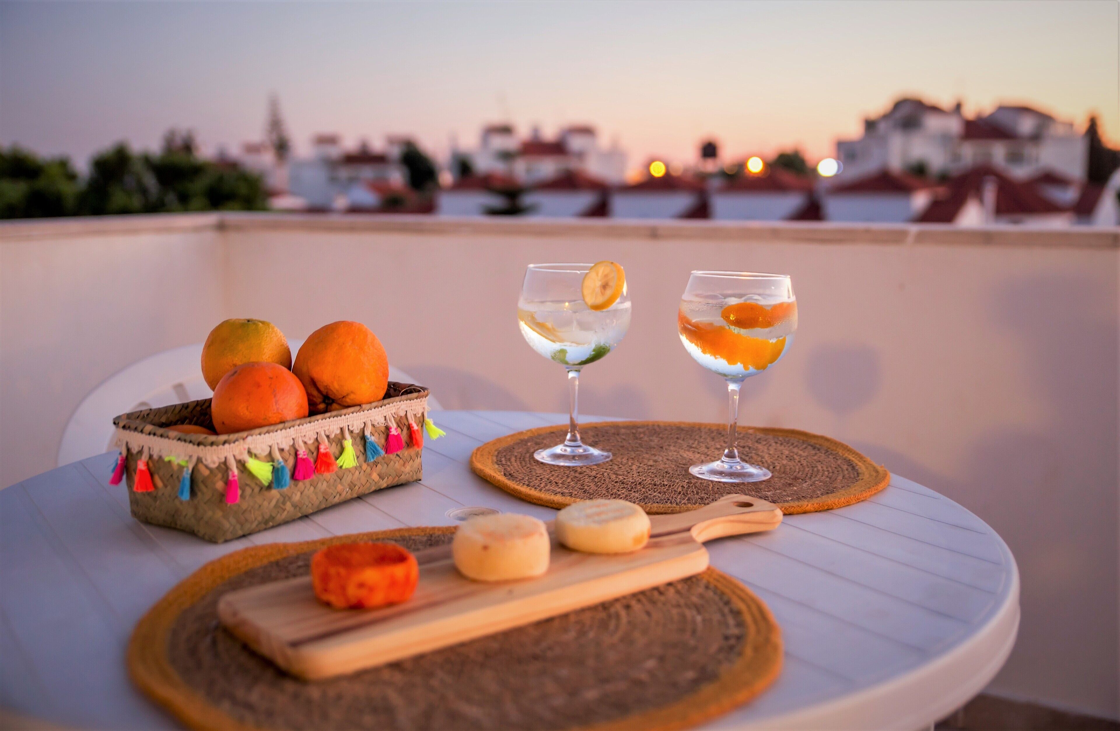 Una mesa con una cesta de naranjas, dos vasos de una bebida cítrica y queso sobre un mantel blanco, con vista a la ciudad de fondo.