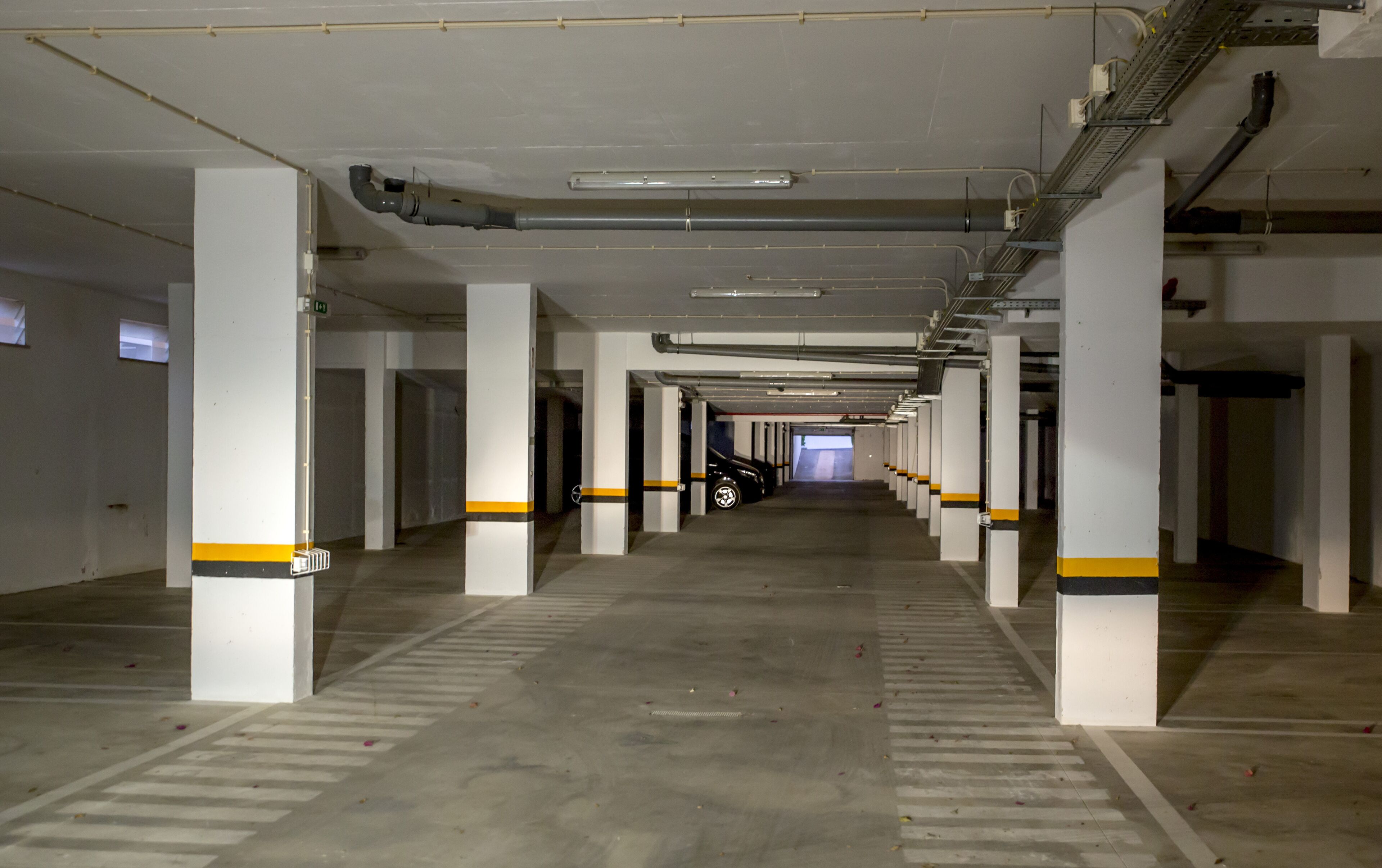 Un long parking souterrain, vide, avec des piliers blancs et des rayures jaunes, un sol en béton et des tuyaux apparents au plafond.