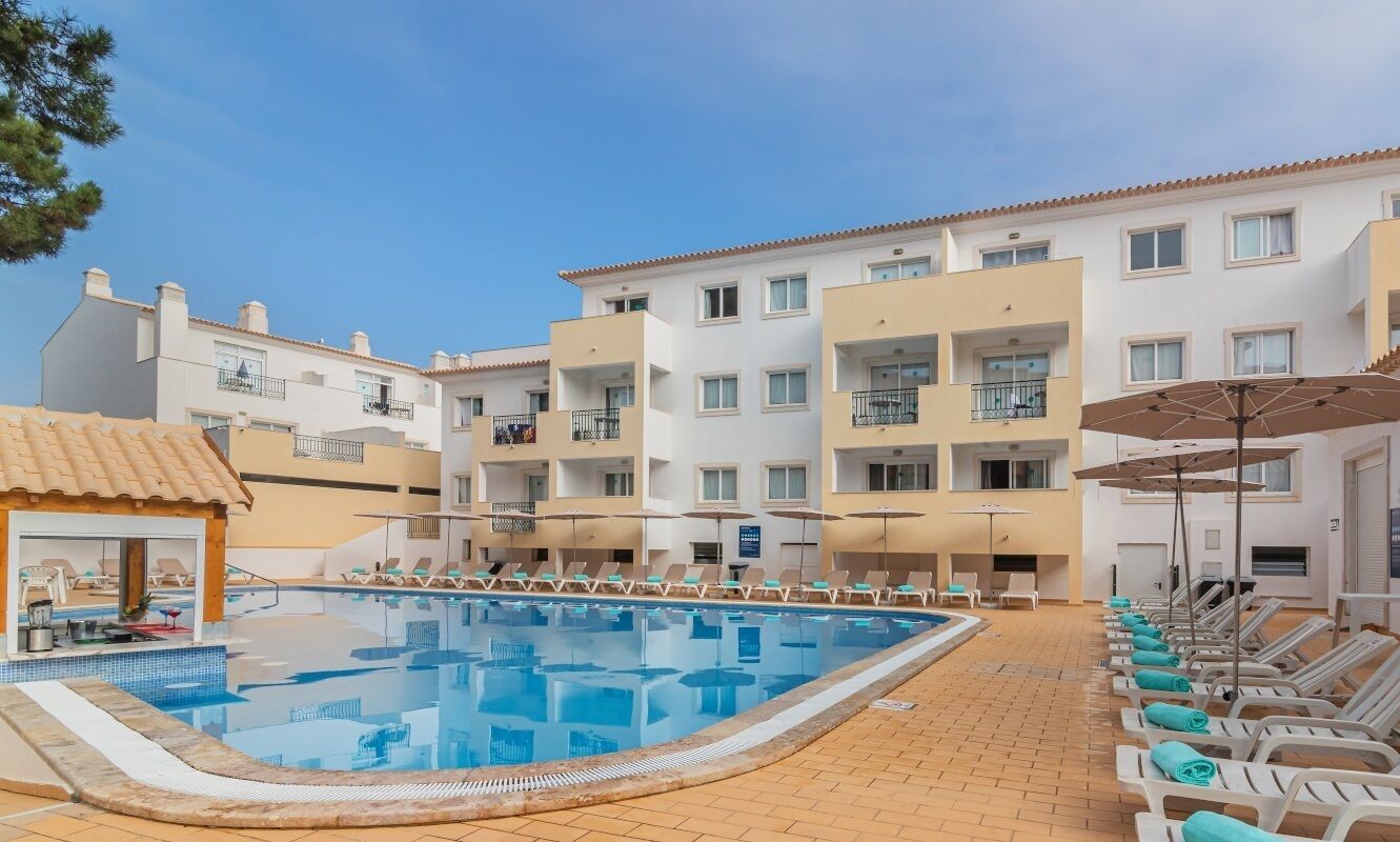 A hotel with a swimming pool, sun loungers, and a covered area with umbrellas.