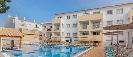 A hotel with a swimming pool, sun loungers, and a covered area with umbrellas.
