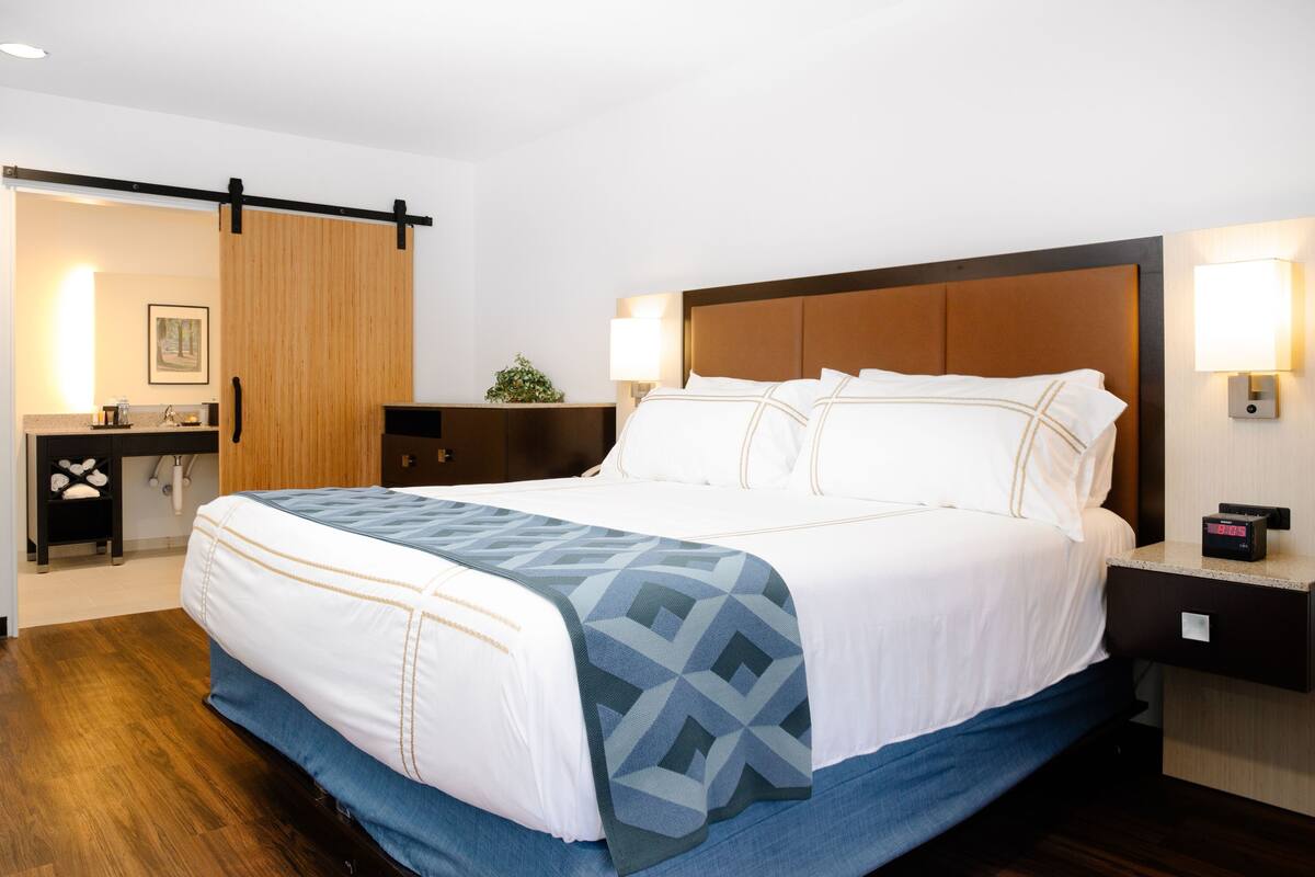 A hotel room with a large bed, a wooden headboard, bedside lamps, a nightstand, and a desk with a plant.