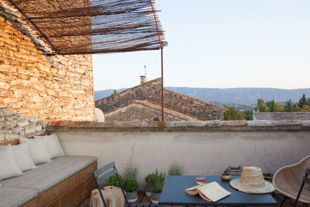 Une terrasse sur le toit, avec un mur en pierre, un banc rembourré, une table sur laquelle se trouvent un chapeau et un livre, ainsi que des plantes en pot.