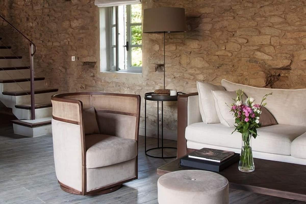 Un salon avec un mur en pierre, un canapé, un fauteuil, une table basse, un lampadaire et un escalier.